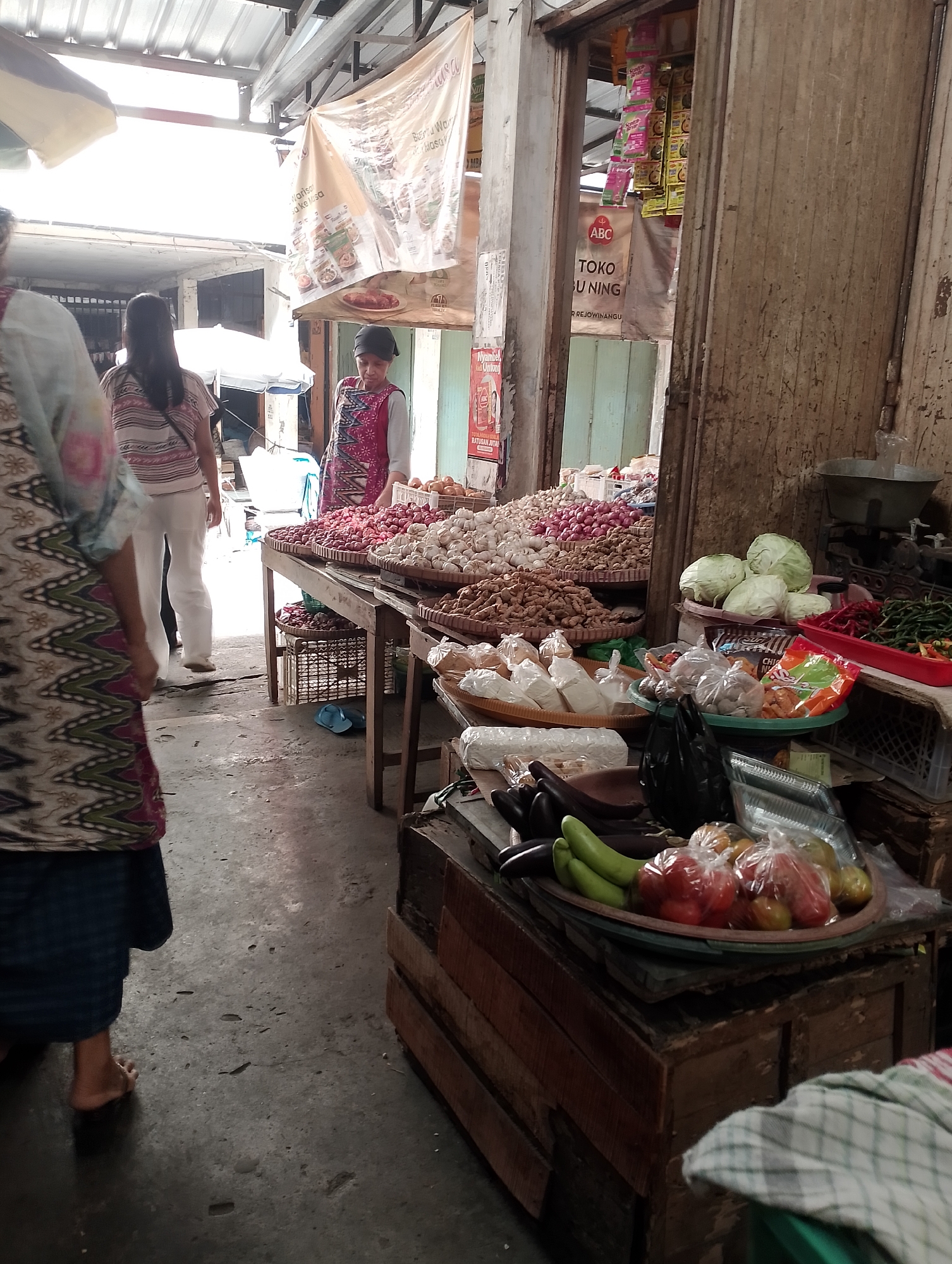 An outdoor market stall in Magelang, Indonesia, displays various fresh produce. A woman in a patterned apron stands behind a table laden with baskets of red onions and garlic. In the foreground, a customer, partially visible from the side, is looking at the produce. The stall is set up on a concrete floor beneath a metal roof structure. Produce such as cabbage, tomatoes, chilies, and eggplants are also displayed on wooden crates and trays. Packaged goods and seasonings are visible in the background.