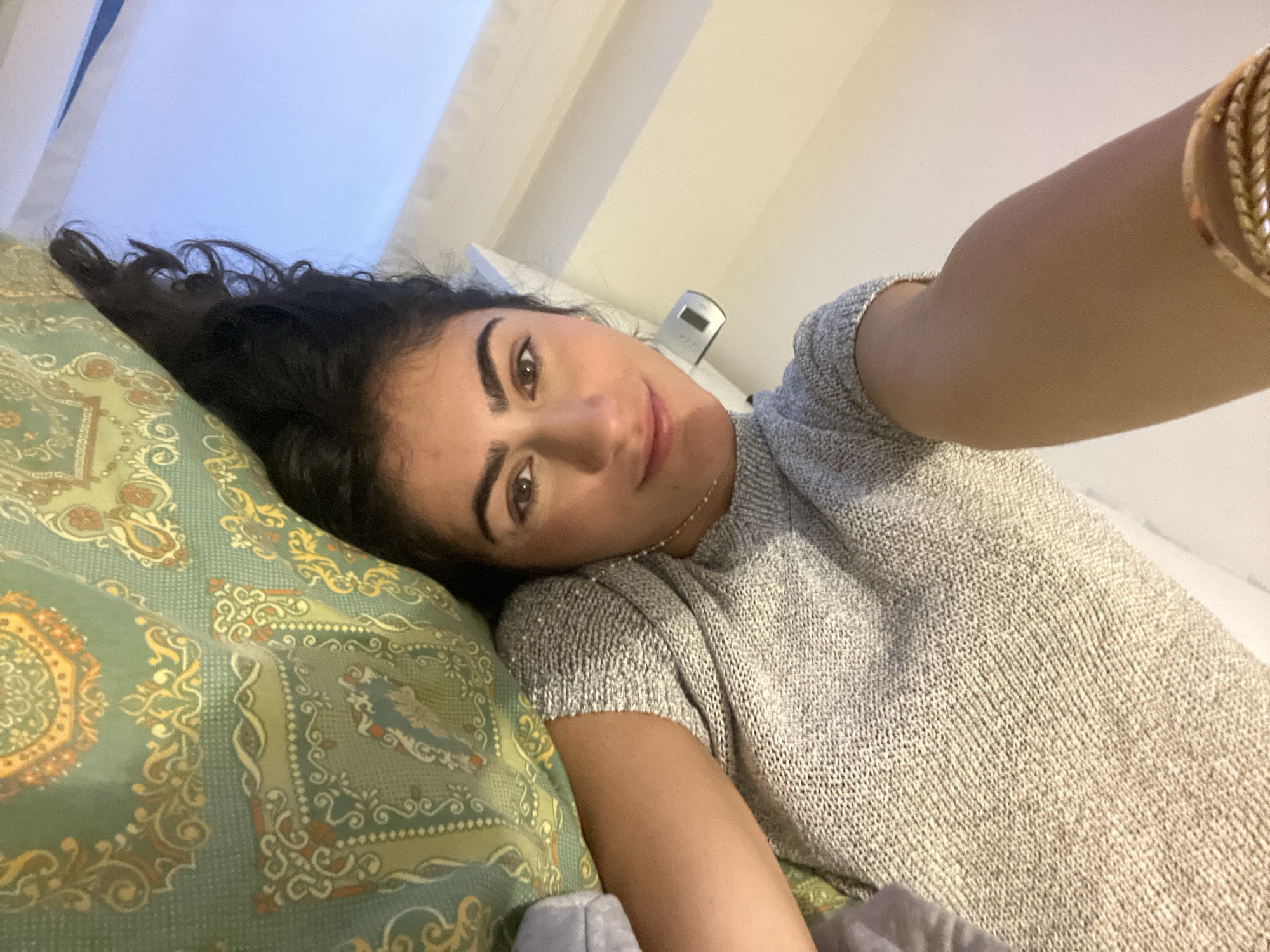 A selfie captures a young woman lying down in an indoor setting, likely a bedroom or living room in Milan, Italy. She has dark, wavy hair and expressive eyebrows, looking directly at the camera with a soft, relaxed smile. She is wearing a light grey, short-sleeved top with a visible knit texture. On her right wrist, she sports several gold-colored bracelets, one appearing braided. Her head rests on a patterned pillow or cushion featuring intricate designs in shades of green, gold, and brown/rust. In the background, a light-colored wall is visible, along with a small digital clock or electronic device on a surface. The lighting is soft and ambient, suggesting it might be late afternoon or evening. The overall mood is calm and comfortable. There is no visible text in the image.