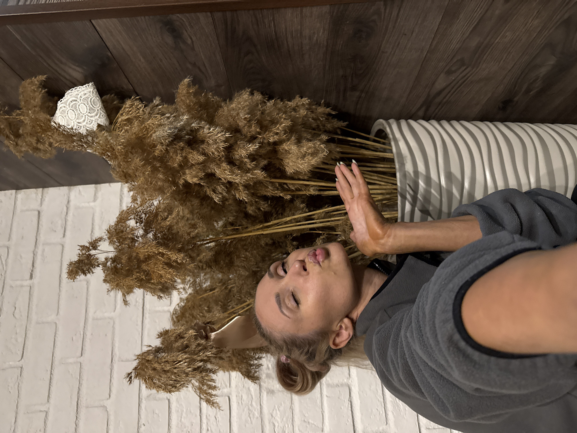 Location: Chaikovsky, Russia.
The image is an indoor photograph featuring a single woman posing for a selfie. The woman, an adult, is visible from the upper torso, angled to the left, with her head tilted slightly upward. She has blonde hair styled in two small buns and is wearing a charcoal gray fleece jacket with a visible black zipper. Her eyes are closed, and her lips are puckered in a kiss gesture directed towards the camera. Her right hand is extended forward, palm open and slightly cupped, displaying fingernails with white tips.
The background consists primarily of a dense arrangement of tall, dried, light brown, feathery plants, consistent with pampas grass, contained within a tall, white, cylindrical vase featuring a pronounced horizontal wavy or ribbed texture. The vase rests on the floor. To the left of the plants, a white brick wall is visible. To the right, a dark brown wooden paneled wall, composed of horizontal planks, serves as the background. A small, folded piece of white lace with a circular pattern is suspended from one of the dried plant stalks near the wooden wall. A beige-colored shoe, possibly a high-heeled pump, is partially visible amidst the plants above and behind the woman's head.