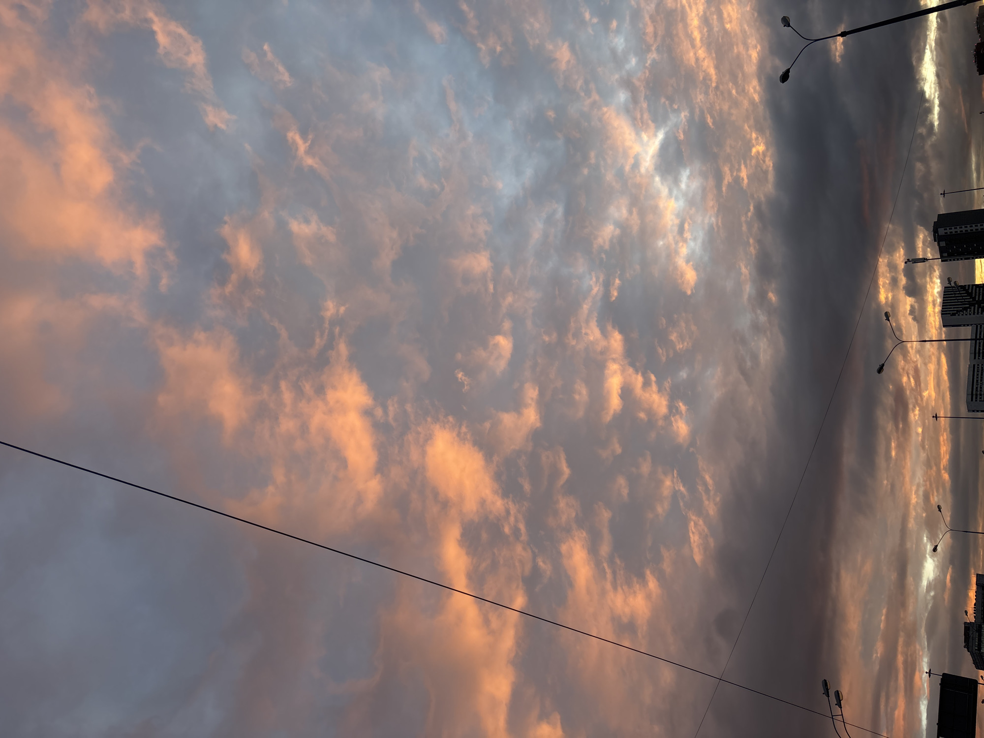 The image shows a sky filled with clouds illuminated by the setting sun, exhibiting hues of pink and grey. The scene is framed by overhead power lines. In the background, city infrastructure including buildings and streetlights are visible. Based on the style of architecture and streetlight design, this could possibly be the city of Moscow, Russia.
