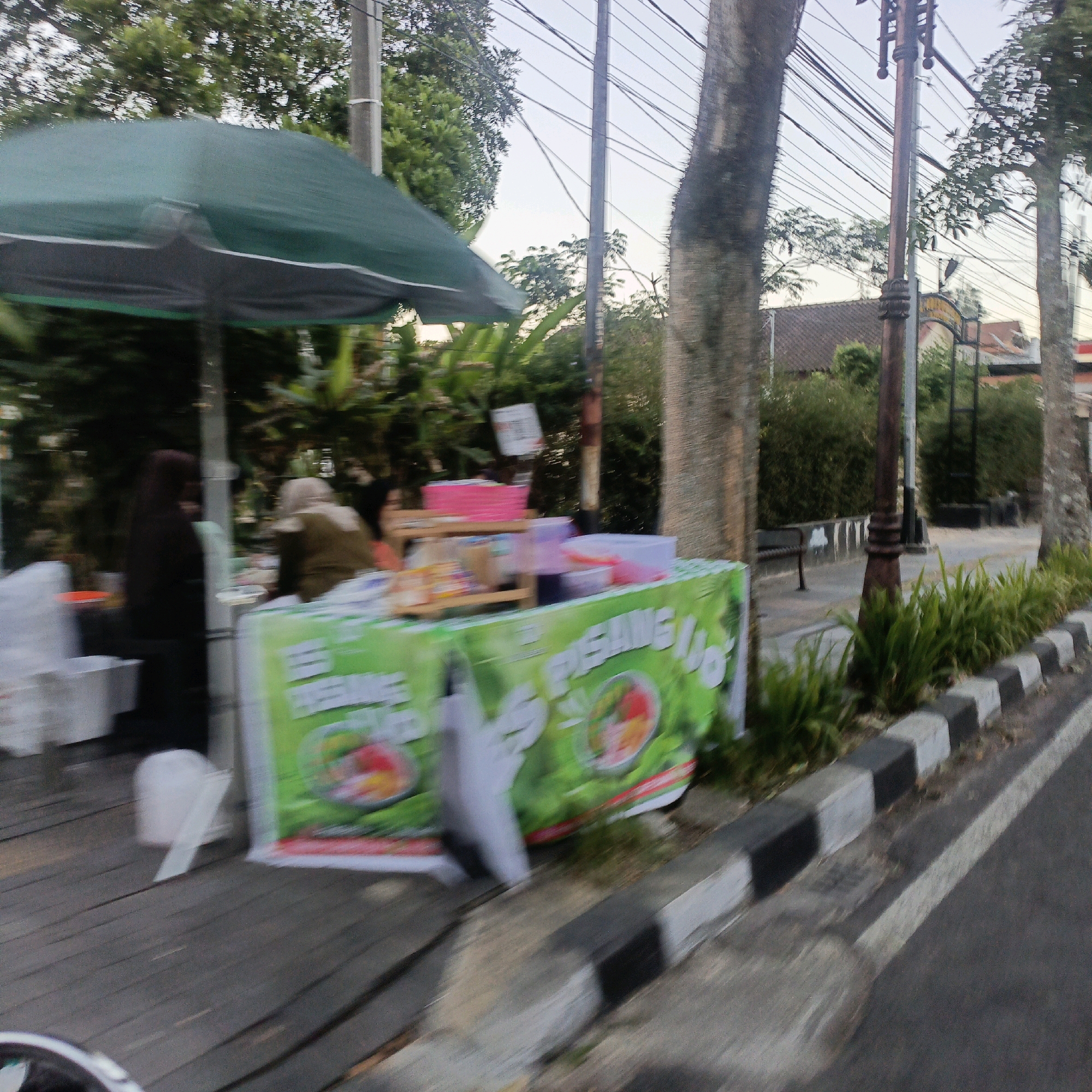 The image captures a street-level view of a food stall in Salatiga, Indonesia. The stall, draped with a green banner featuring text and images of fruit bowls, is set up on a wooden platform. To the left, a green umbrella provides shade over part of the setup. Visible on the stall are stacked pink containers and various food items in transparent containers. Two individuals are partially visible behind the stall; one wearing a dark headscarf and another in a light-colored hijab. In the background, trees and power lines suggest an outdoor, possibly roadside, setting. The foreground shows a paved sidewalk with a curb, indicating this is likely along a street. The overall scene is slightly blurred, suggesting movement, perhaps from a vehicle.