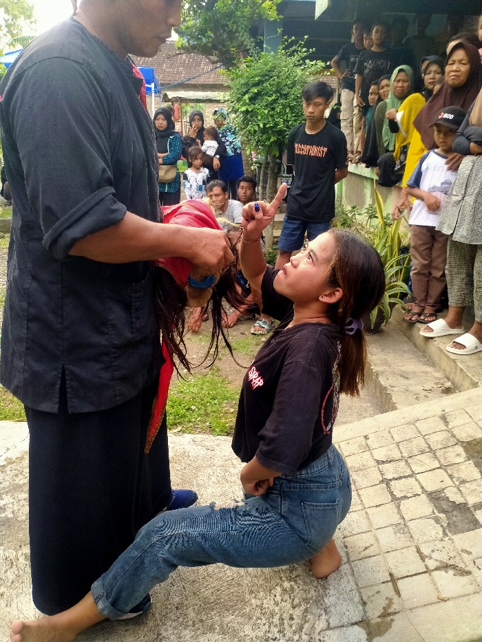 The image depicts an outdoor scene in Jenggrik, Indonesia, featuring two primary figures engaged in an interaction before a crowd of spectators. A large man, facing right and clad in a black long-sleeved shirt and black trousers, stands with his bare feet on an uneven concrete surface. He holds a traditional, ornate object, possibly a mask or headdress with red fabric and dark hair, upside down in his hands. Before him, a smaller woman is kneeling on a patterned concrete slab, barefoot, wearing a black t-shirt with red lettering and ripped blue jeans. She looks upwards towards the object held by the man, her mouth slightly open, and her right index finger, painted with blue nail polish, is extended upwards and pointed towards the object. Her dark hair is pulled back with a purple tie.
Approximately twenty spectators, a mix of adults and children in varied casual attire, observe from behind the two central figures. They stand on a slightly elevated concrete area and dirt ground. The background features green foliage from bushes and trees, and a residential building with a tiled roof is partially visible. The scene is illuminated by natural daylight.