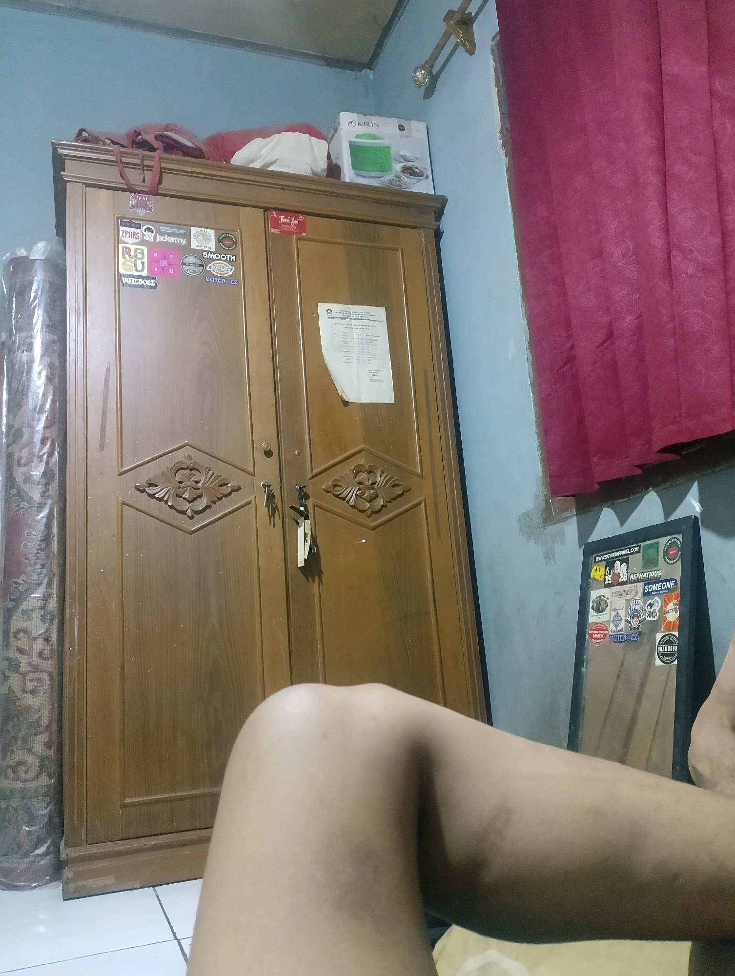 In the city of Ciomas, Indonesia, a wooden wardrobe stands against a light blue wall, adorned with various stickers. On top of the wardrobe, a red cloth bag and a white sheet are visible. To the right of the wardrobe, a section of a red curtain hangs, and a mirror with numerous stickers is propped against the wall. In the foreground, a person's bare leg is visible.