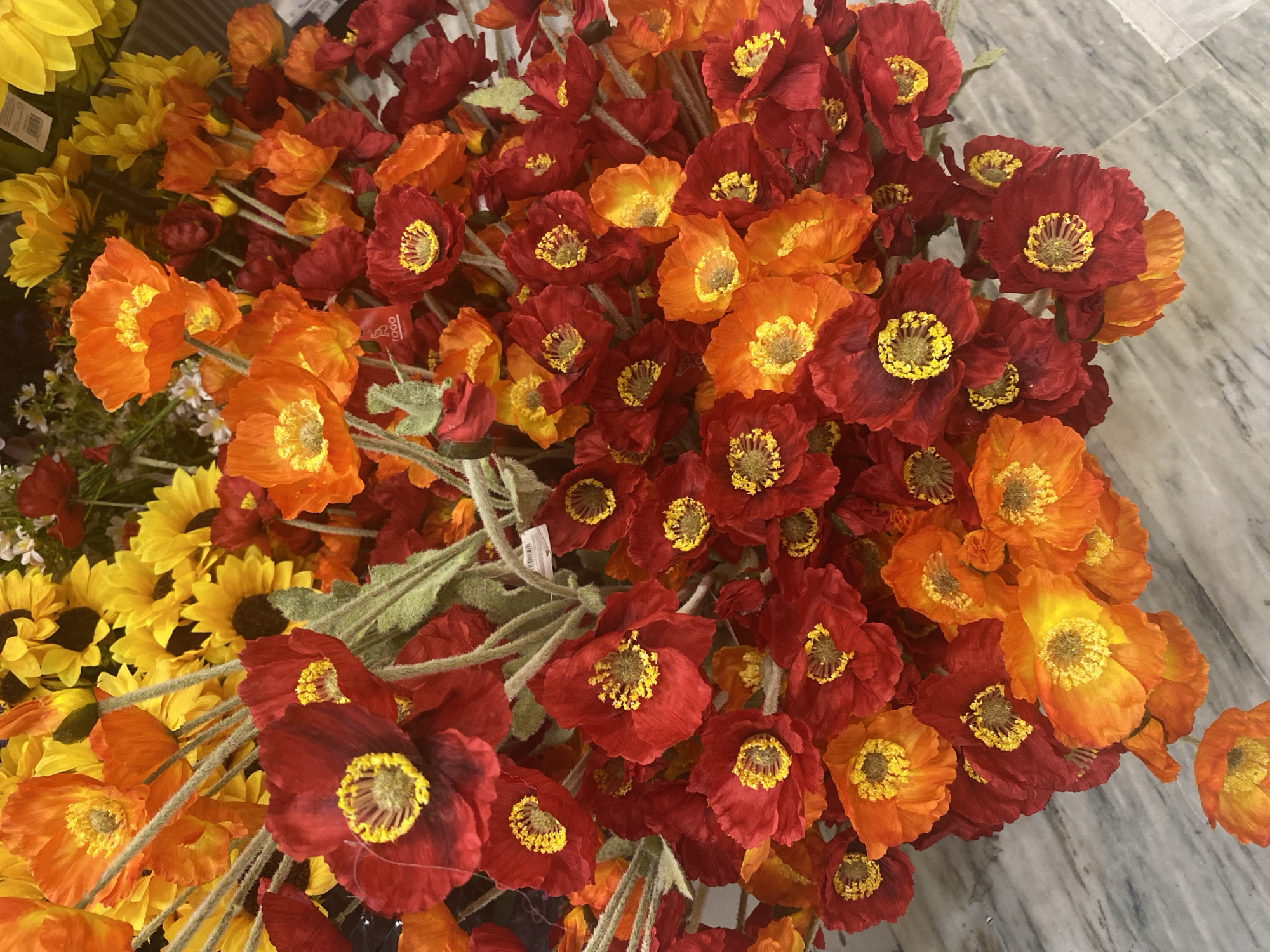 A close-up overhead shot features a large bouquet of artificial poppies in varying shades of red and orange. The flowers have delicate, crinkled petals and prominent yellow stamens. They are arranged closely together, with some stems and fuzzy green foliage visible. The backdrop is a light gray marble surface with subtle veining. No people or specific landmarks are present, making it impossible to determine a location within Konya, Turkey.