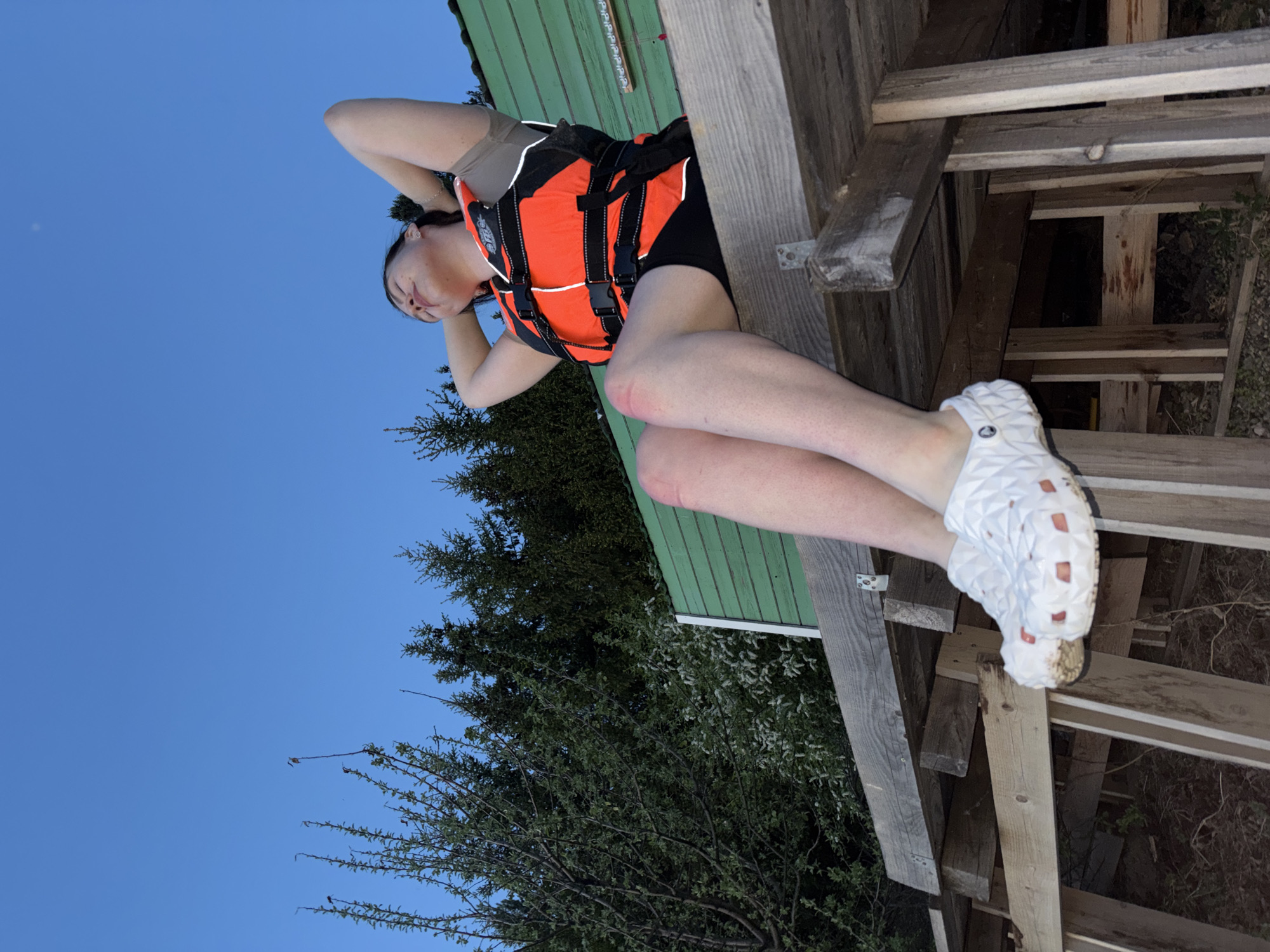 A woman is lying on a wooden structure near Gora, Russia. She is wearing a life vest and white Crocs. A green building and trees are visible behind her. The sky is blue.