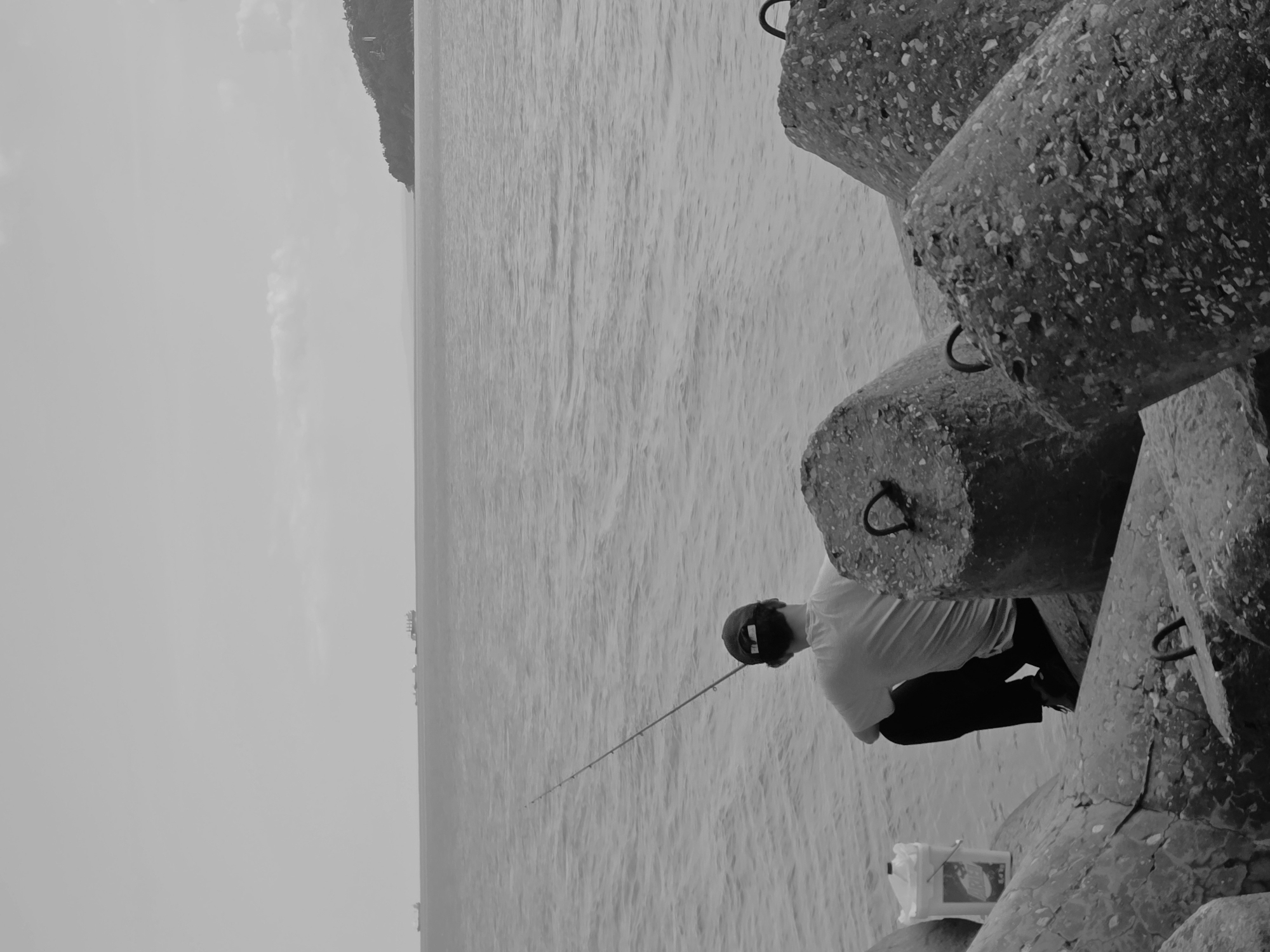 A grayscale photograph depicts a man fishing from a breakwater in Varna, Bulgaria. The man, wearing a baseball cap, a light-colored t-shirt, and dark trousers, is seated on the large concrete tetrapod breakwater. He holds a fishing rod that extends out over the water. A white bucket sits next to him. The background features the sea, a distant coastline, and a hazy sky.