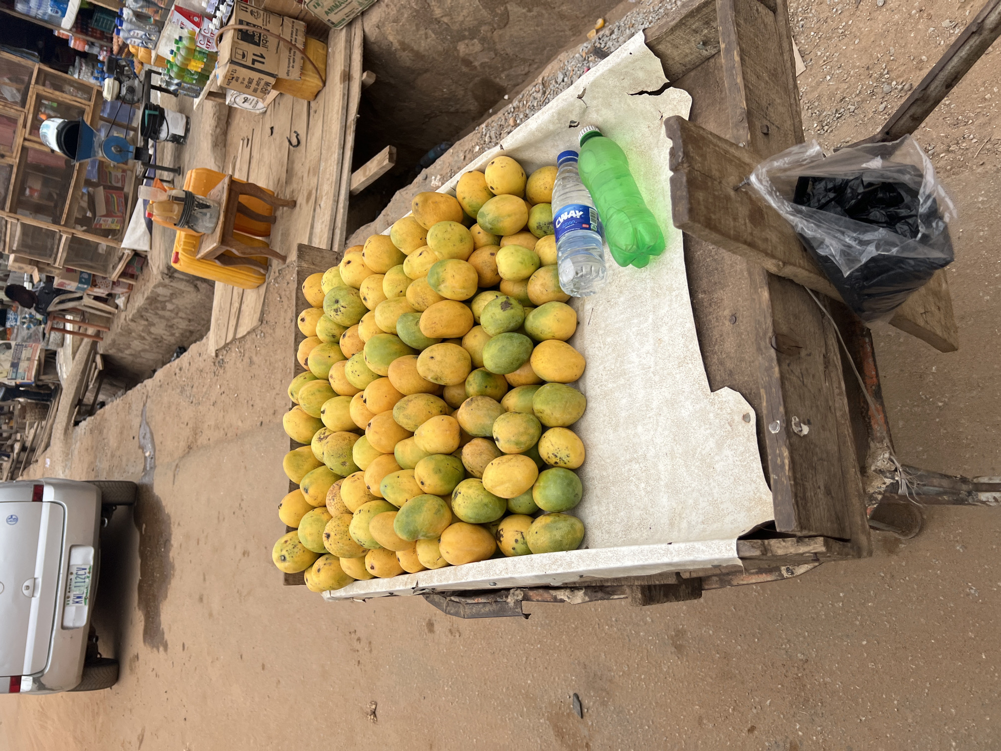 The image depicts a roadside scene in Abuja, Nigeria, focusing on a handcart laden with a large pile of mangoes. The mangoes, varying in color from yellow to green, are stacked high on a white sheet-covered surface within the cart. A bottle of water (brand name AWAY) and a green plastic bottle sit among the fruit. A black plastic bag is attached to the cart.
In the background, a partially visible silver car with license plate "KWL-112CV" is parked near the cart. A vendor's stall with shelves displaying various products is located near the vehicle. The environment is dusty, and the stall's surroundings suggest a bustling commercial street or market.