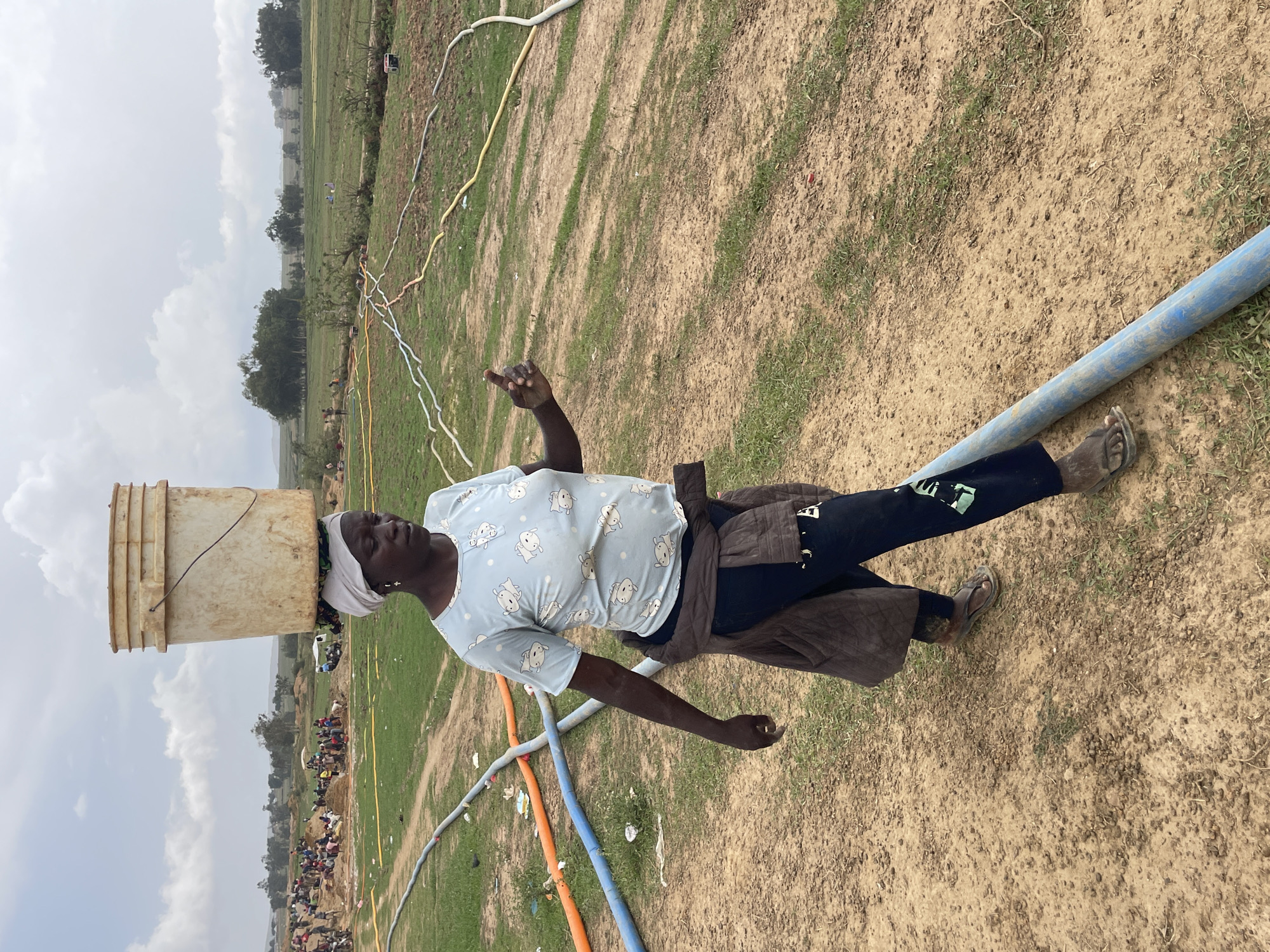 In an outdoor scene in Durumi, Nigeria, an individual stands on a dirt and grassy area. They balance a tan bucket on their head, and are dressed in a light blue t-shirt with a cartoon pattern, dark pants, and sandals. A white head covering is visible. The person is pointing. In the background, numerous people are clustered, and several hoses lay across the ground. The sky is overcast.