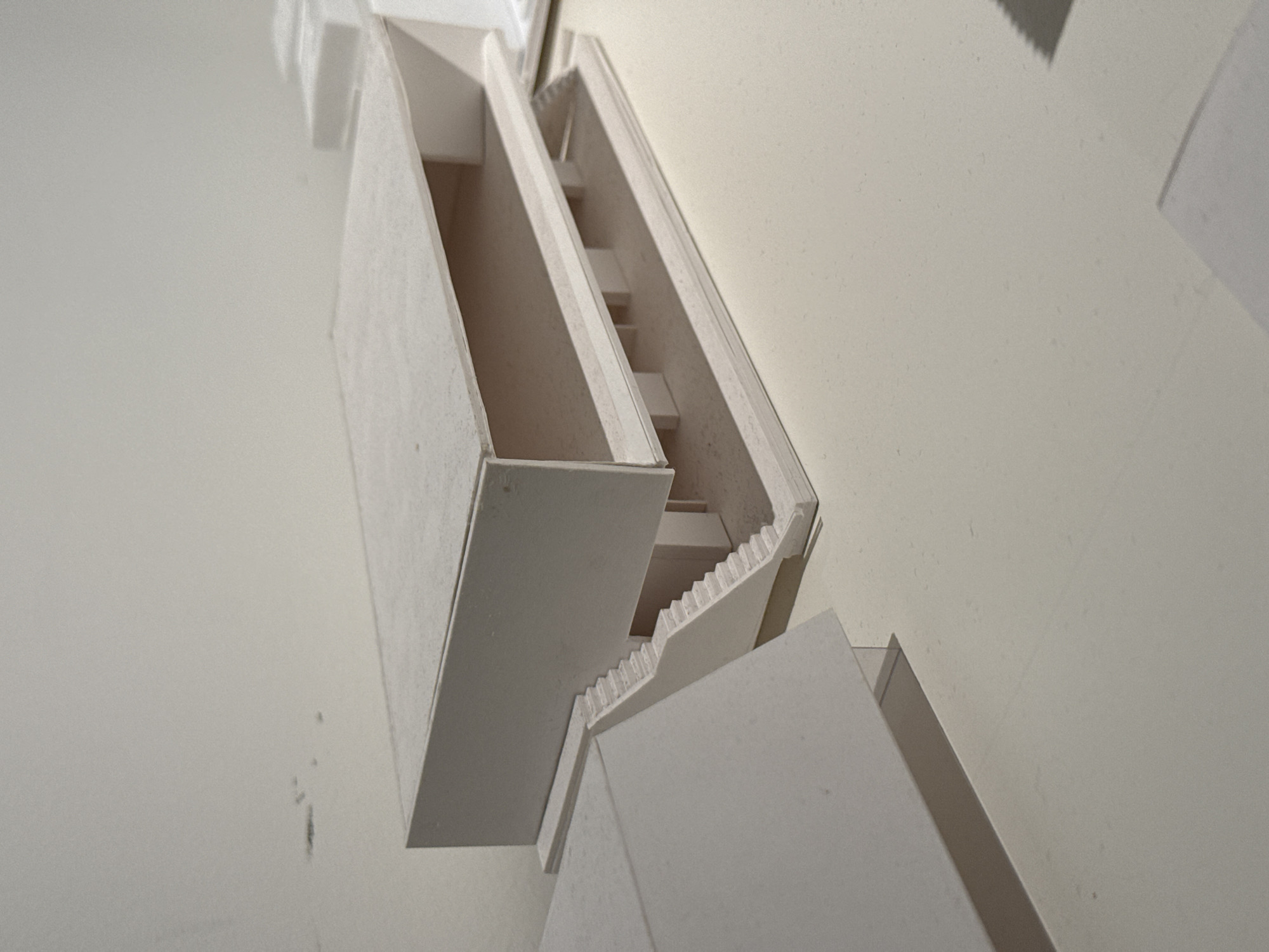 Location: Milan, Italy.
The image presents a detailed view of a static architectural model, predominantly white or light grey, displayed on a smooth, light-colored surface. The central model is a multi-level, rectilinear structure featuring an upper, solid rectangular block resting on a lower, open-plan section. The lower section includes a base slab, an intermediate floor supported by several slender vertical columns, and an open space beneath the upper block. A prominent element is a staircase ascending from the left side of the model's base, leading to the intermediate level, characterized by individual steps and a solid side banister. To the immediate left foreground, a portion of another white, angular architectural element is visible, casting a distinct shadow on the display surface. In the background, to the right of the central model, faint outlines of additional white rectangular forms are discernible, suggesting other architectural models or components arranged in a row. The display surface extends into a plain, light-colored wall in the background, creating a clean, minimalist environment. The overall lighting is soft and even, casting subtle shadows that define the geometric planes and depth of the model. No individuals, specific actions, or interactions are present in the scene.