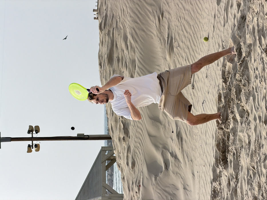 A lone adult male, appearing Caucasian with short brown hair and a light beard, is depicted barefoot on a sandy beach. He wears a white short-sleeved t-shirt and tan shorts. He is captured in an athletic stance, knees bent, holding a bright neon-green paddle above his head, actively engaging in the sport of matkot. His gaze is fixed on a small, dark spherical ball suspended in mid-air to his left, which he is poised to strike. A second yellow-green ball rests on the sand to his lower right. The immediate environment consists of undulating sand dunes. In the background, the Mediterranean Sea is visible, featuring a rocky breakwater. A wooden pergola with a slatted roof and the number "3" on a post is partially visible on the far left, behind the dunes. A tall, dark utility pole equipped with four horn-shaped speakers or lights stands against a clear, light grey sky in the central background. A small, dark bird is in flight in the upper right portion of the frame. The scene is located on a beach in the vicinity of Tel Aviv Port, Tel Aviv, Israel.