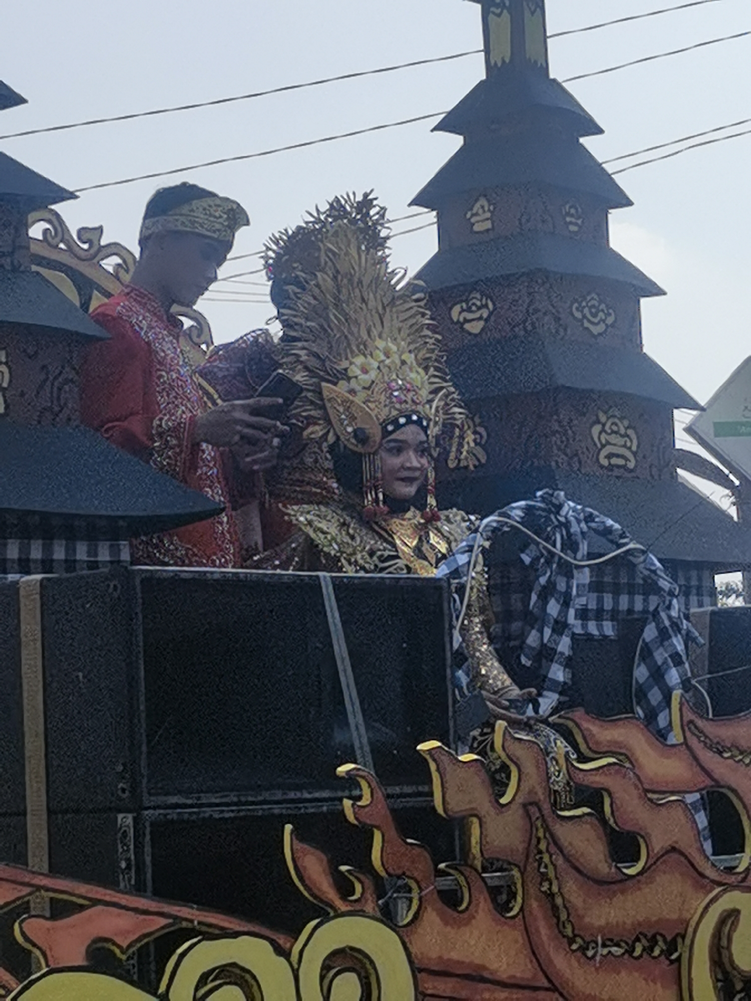 The image depicts a parade float in Blora, Indonesia, featuring two individuals in traditional attire. On the left, a person wearing a red garment with intricate gold embroidery and a golden headpiece is visible. This individual is looking downwards and holding a black smartphone. To the right, a person adorned in a highly ornate golden headdress and matching costume is seated. This individual is smiling and looking to the side. Behind them, a multi-tiered, dark-colored structure with decorative motifs resembling flowers is part of the float's design. A black and white checkered sarong is draped over an object on the right side of the float, and the foreground shows a colorful, flame-like decoration in shades of orange and yellow, suggesting a festive or ceremonial occasion. The background is a hazy sky, and power lines are visible stretching across the upper portion of the frame. The float appears to be stationary, possibly during a parade or procession.