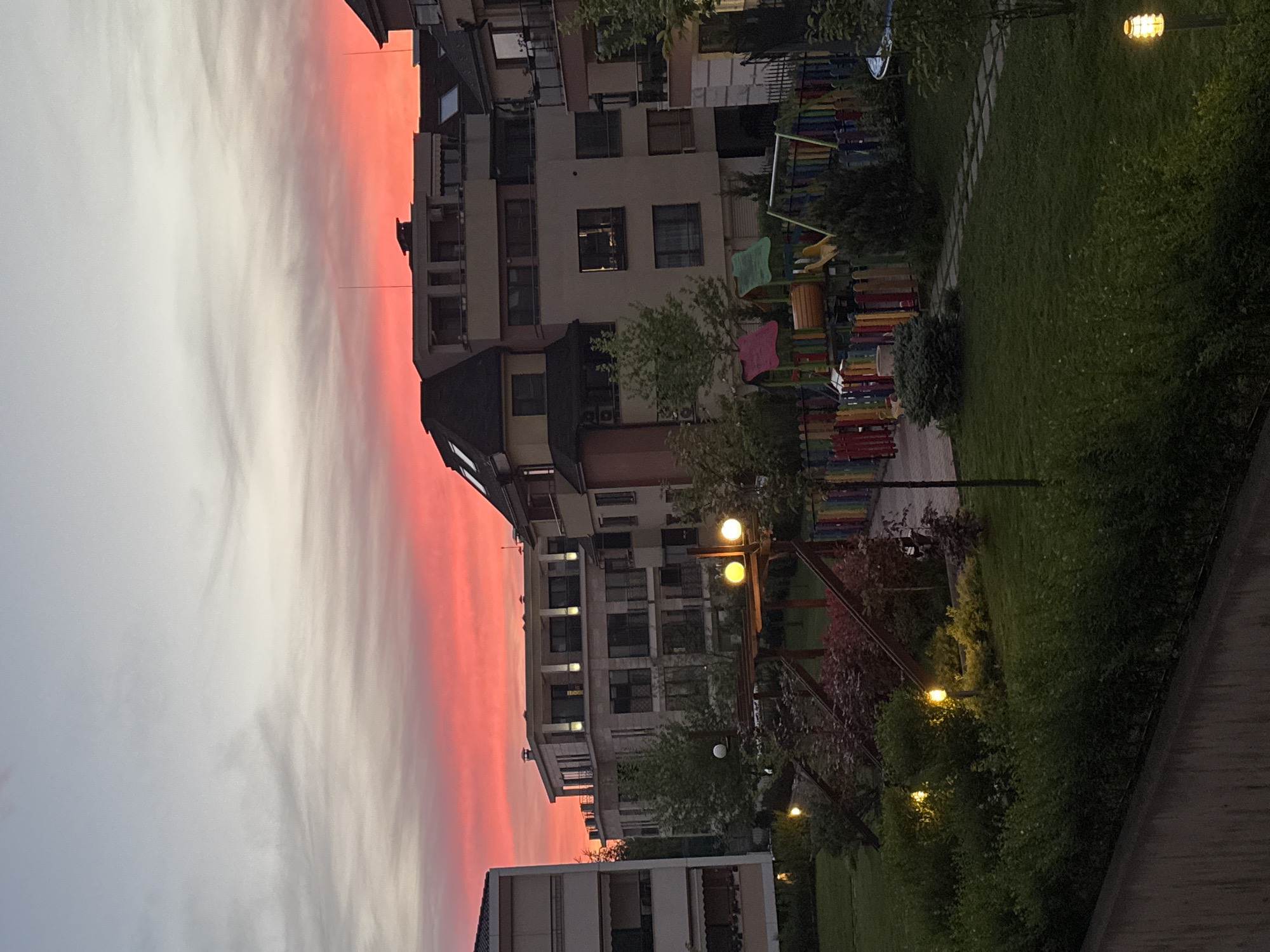 Captured from a tilted perspective, the scene presents a cityscape at dusk, likely in Sofia, Bulgaria. The sky transitions from a fiery red hue near the horizon to a paler shade above, suggesting a recent sunset.
A multi-story building dominates the view. The building features multiple windows and balconies. Adjacent to the building is a playground area with colorful equipment like swings, slides, and climbing structures. The area is illuminated by multiple light sources, including lampposts.
The foreground is defined by lush greenery. A pathway, likely part of a public park, borders the green space. A section of a concrete structure or wall is visible at the bottom.