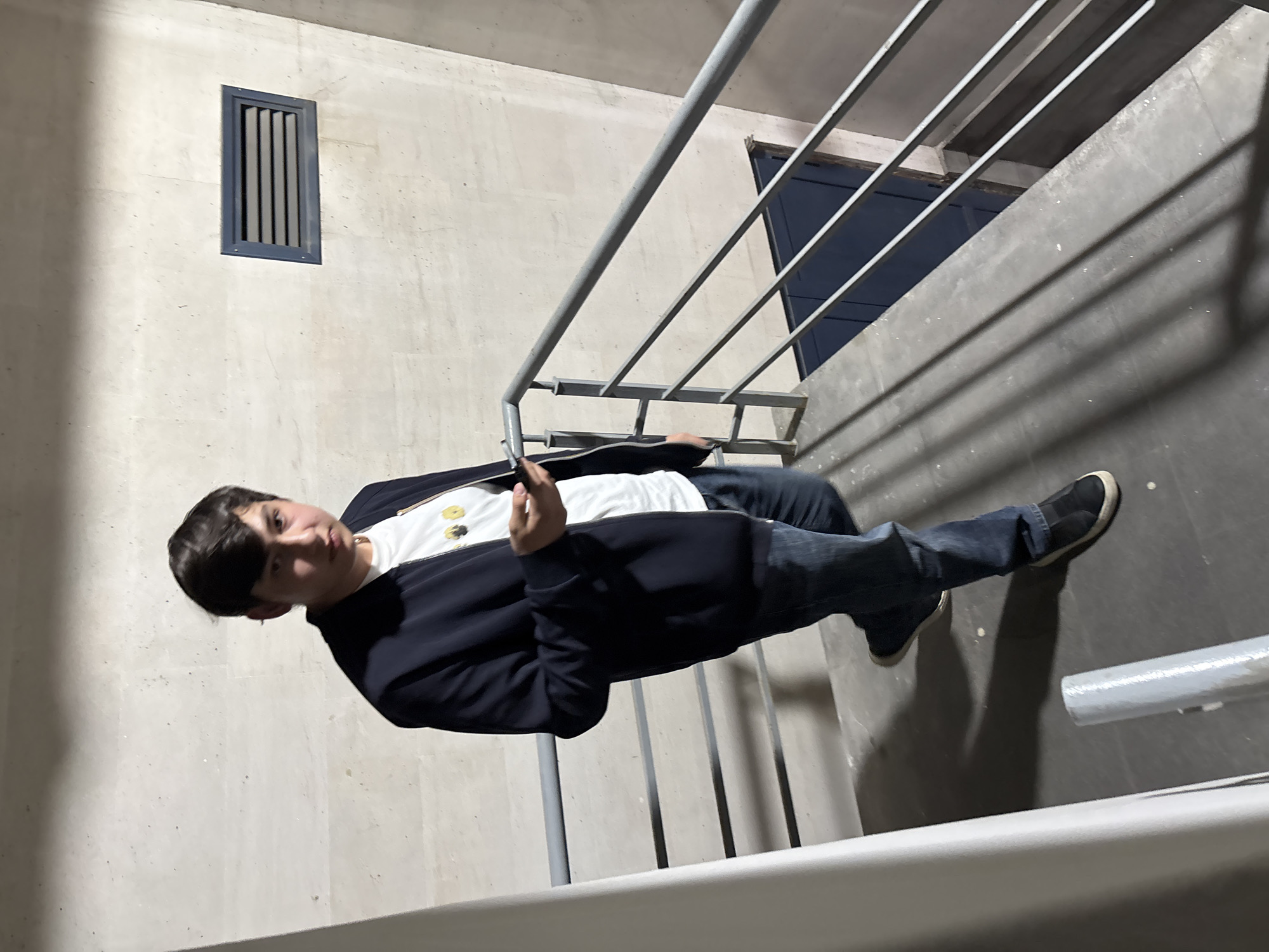 A young male is captured standing on a concrete stairwell in what appears to be an indoor or covered area, possibly in Yerevan, Armenia. The original image is rotated, showing the person vertically aligned with the stairs.
The individual, seen from the waist up in the main frame, wears a dark jacket over a white t-shirt that has some dark circular details or buttons on the chest. He also wears blue jeans and dark slip-on shoes. His hair is short and dark, and his expression is neutral, looking directly towards the viewer. His right hand is slightly raised, with the palm facing forward, in a gesture that is not entirely clear but might be acknowledging the camera or holding something unseen.
The setting consists of light-colored, possibly concrete, walls with visible seams or textures. A rectangular dark gray or black vent with vertical slats is positioned high on the wall to the left of the person. A gray metal railing runs alongside the stairs, dividing the frame. The floor of the stairwell is a darker shade of gray concrete, and shadows cast across it suggest directional artificial lighting or indirect natural light. In the background, behind the railing, there's a dark framed opening, which could be a doorway or a window to another section of the building. The overall ambiance is functional and somewhat stark, typical of a utilitarian building or parking garage stairwell. There is no visible text in the image.