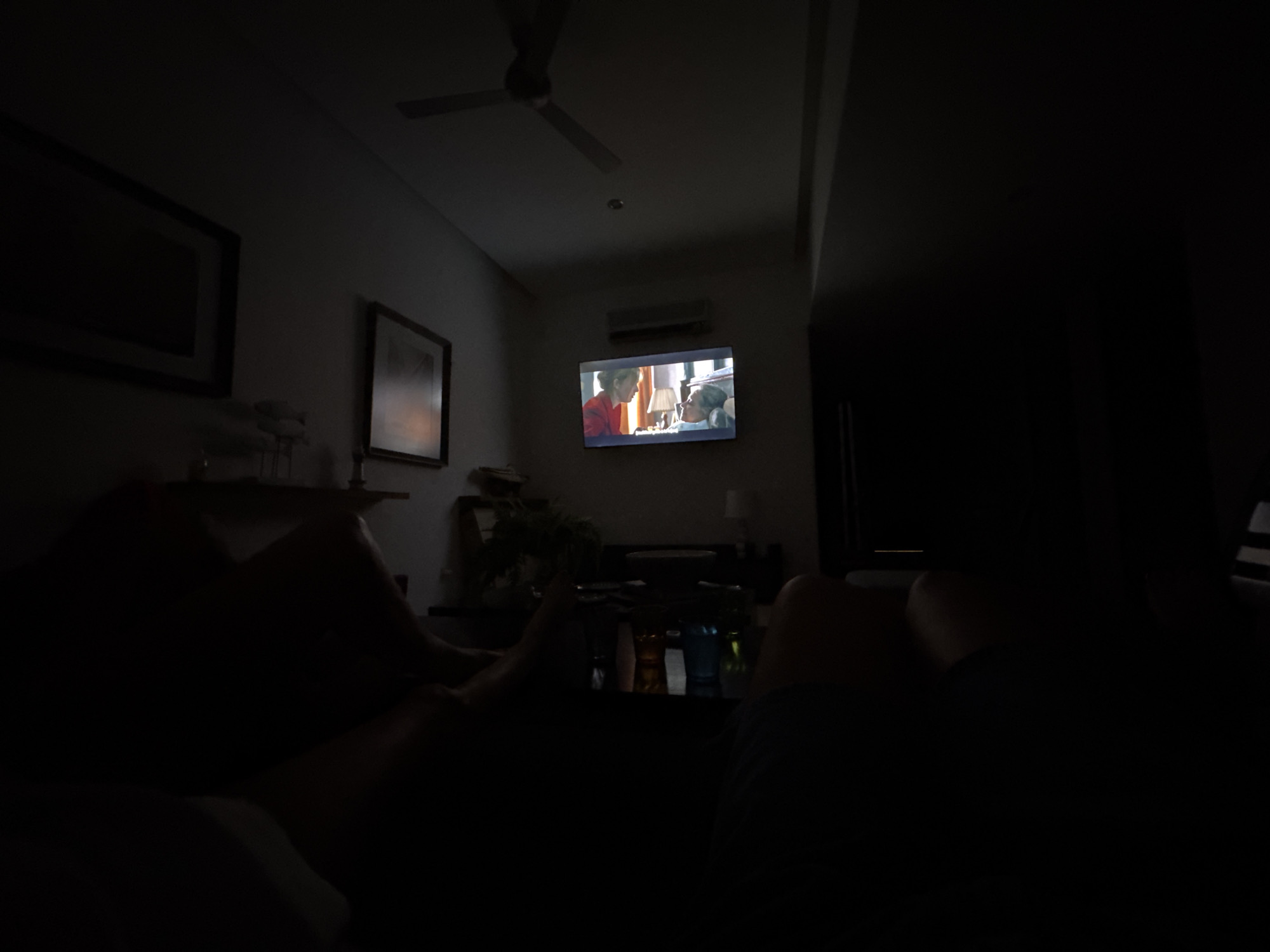 The image shows a dimly lit interior space, possibly a living room, with a television as the focal point displaying a program with two figures. The low lighting obscures most details, but one can discern what appears to be an air conditioning unit mounted above the television. The ceiling fan is visible, and there are frames hanging on the wall. In the foreground, there are vague outlines of what appear to be people sitting, possibly on a couch. A table with objects is located in front of the people.
It's not possible to pinpoint the exact location in Puerto Marqués, Mexico based on this picture.
