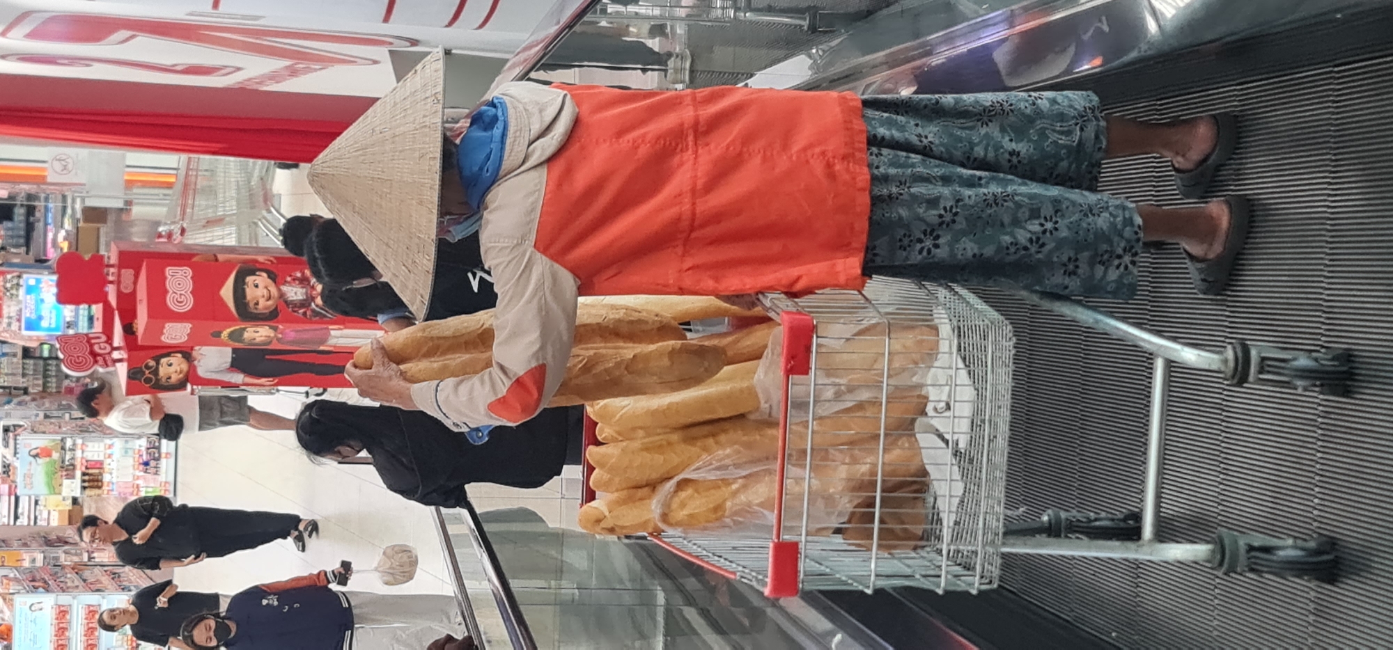 The image depicts a scene inside a brightly lit supermarket or hypermarket, specifically on a moving walkway or escalator designed for shopping carts. The photo is oriented vertically, showing the subjects from a high angle looking down the walkway.
In the foreground, a person is seen wearing a traditional Vietnamese conical hat (nón lá), a vibrant orange jacket over a beige long-sleeved shirt with an orange patch on the elbow, a blue hooded garment underneath, and dark blue trousers with a lighter floral or leaf pattern. They are wearing dark slip-on shoes. This individual is holding a long, golden-brown loaf of bread, akin to a baguette, and is pushing a metal shopping cart. The cart is heavily laden with many similar loaves of bread, some appearing to be wrapped in clear plastic bags.
The moving walkway features a textured, ribbed surface and a metal handrail. In the background, other shoppers are visible on the main store floor, amidst various product displays and shelves. Prominently displayed are large red promotional cutouts featuring cartoon characters. Visible text on these displays includes "Goi" and "GGU," suggesting brand or product names. One of the cartoon characters on the red display also wears a conical hat, echoing the one worn by the main subject. The context suggests a bustling retail environment in Đà Nẵng, Vietnam.