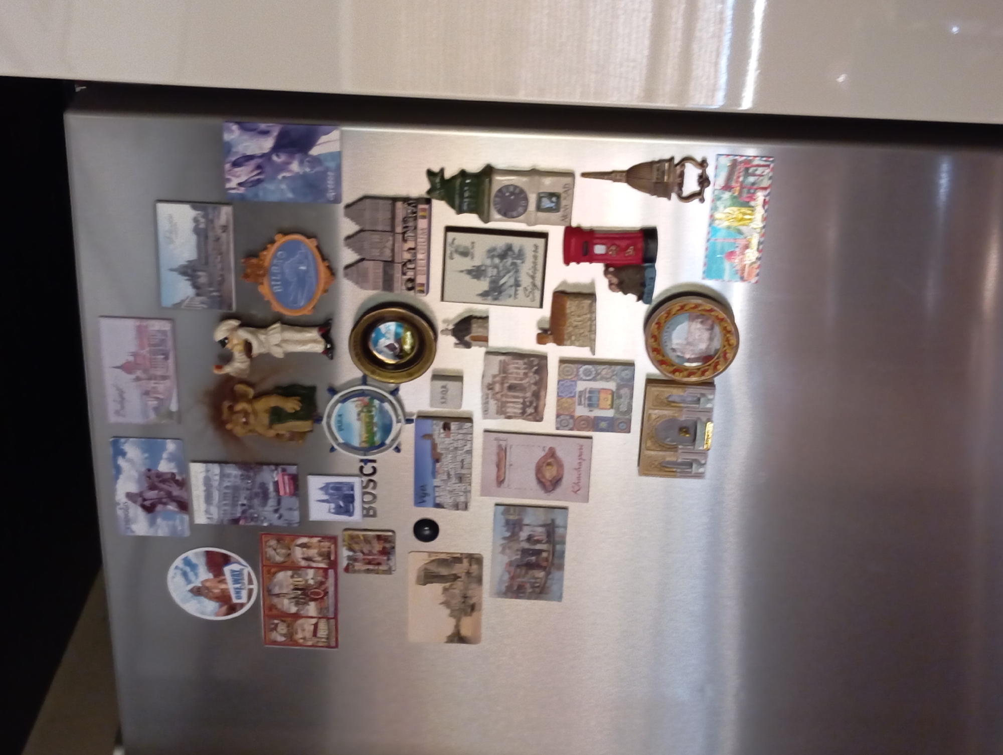 A close-up, eye-level shot shows a collection of novelty magnets adhered to a stainless steel refrigerator door. The magnets are varied in shape and depict miniature scenes, landmarks, and humorous figures, many appearing to be souvenirs from various global locations. Several magnets showcase architectural details and cityscapes, with some suggesting European locations such as Baden, Austria, indicated by a magnet featuring the "Baden" text and a recognizable building. Other magnets include a troll figure, a person in a chef's hat, a red British-style telephone box, and various miniature houses and buildings. The overall impression is of a well-traveled individual with a penchant for collecting mementos from their journeys.