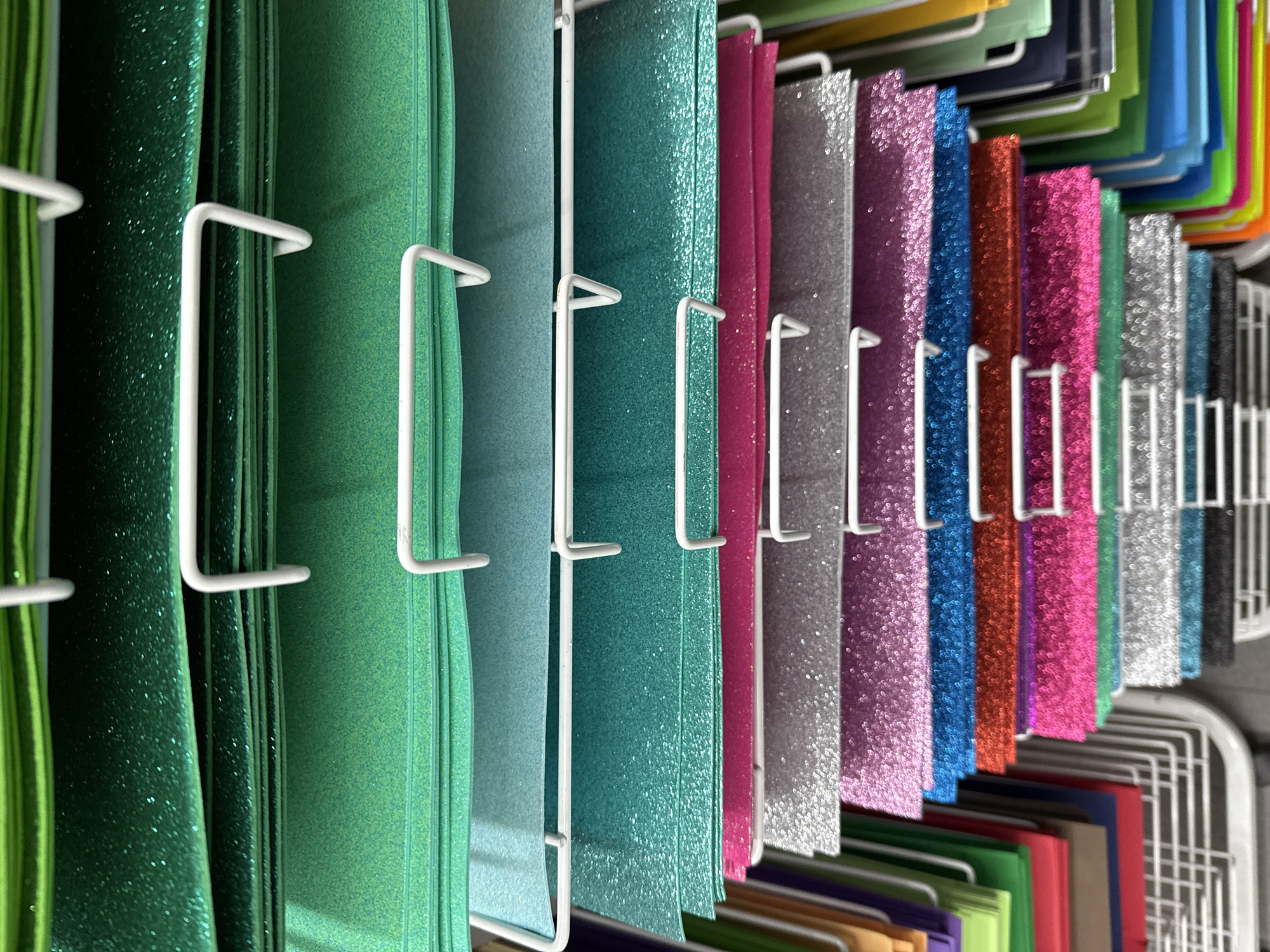 The image depicts rows of glittery craft paper in various colors, including green, teal, pink, silver, purple, blue, and copper. The paper is stacked and held in place by a white wire rack. Other colored papers can be seen along the upper part of the image. The picture looks like it could have been taken at a store. There is no indication that the image was taken in Madrid, Spain. There are no people visible in the image.
