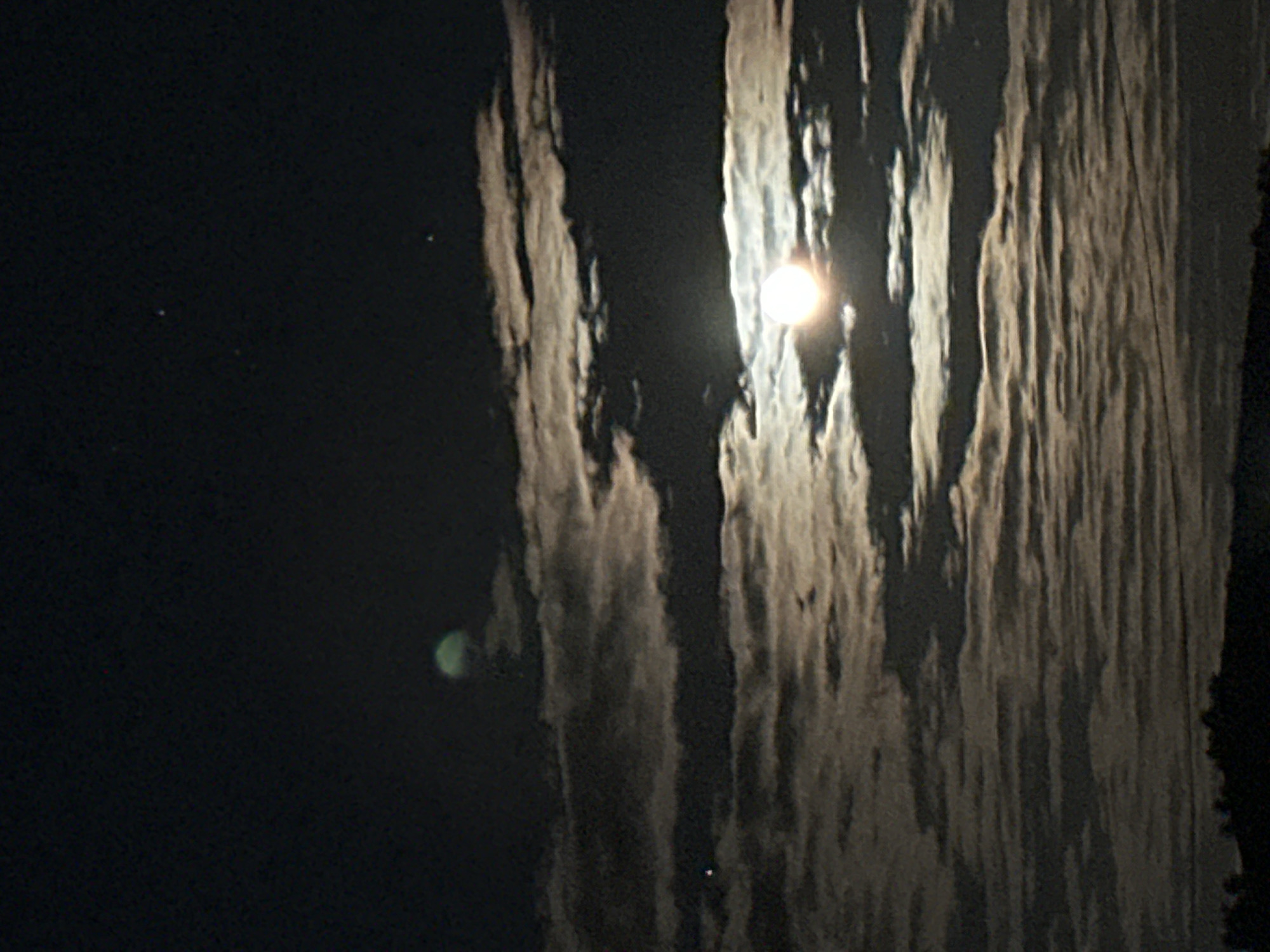 The image shows a night scene in Unknown, Unknown. A bright moon is visible through a vertical band of clouds. The left portion of the image is dark, with a few faint stars and a green artifact from the lens. The right edge shows the silhouetted outline of a tree.