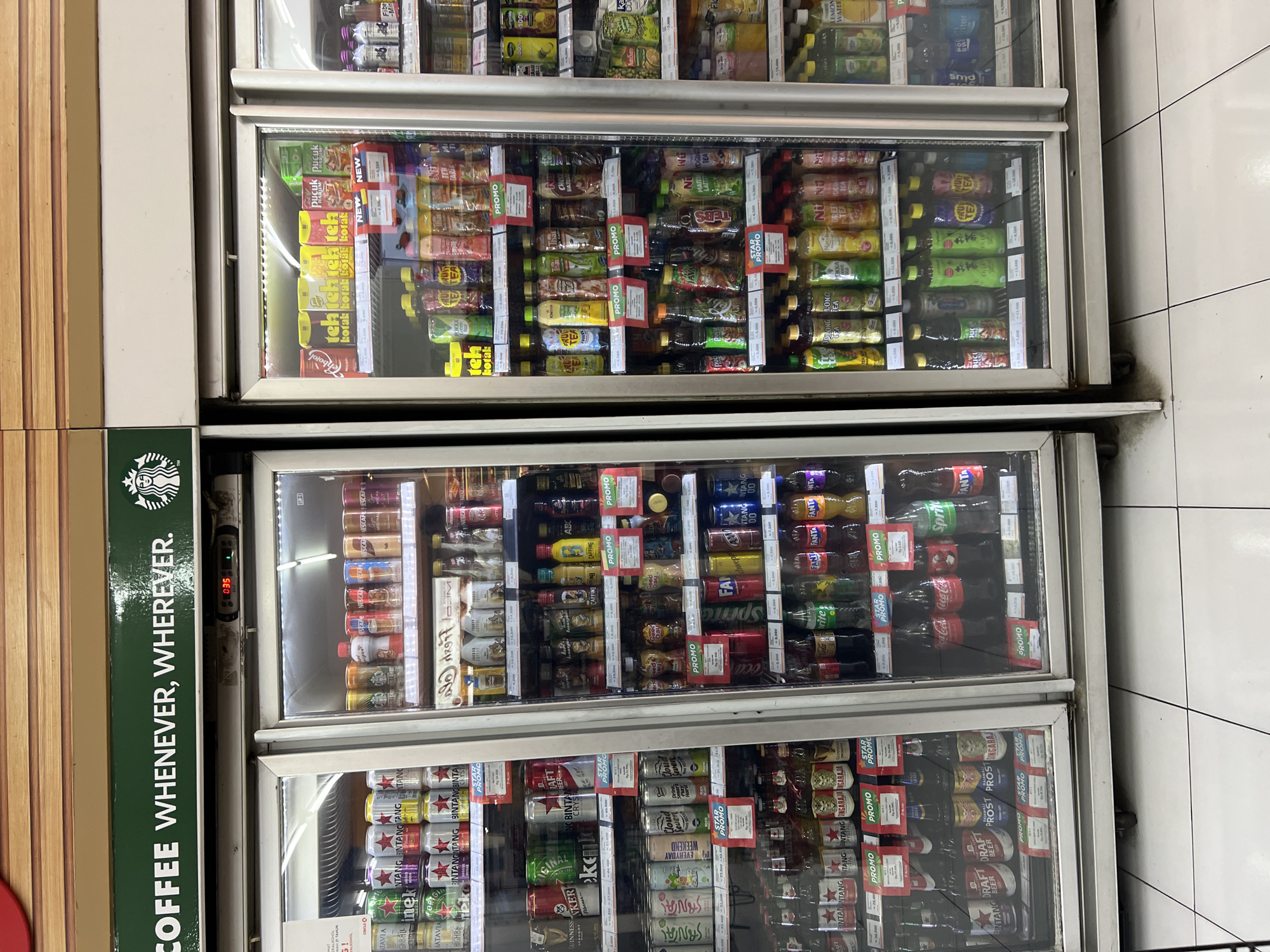 The image shows the interior of a convenience store, likely a 7-Eleven, with multiple refrigerated display cases filled with beverages. Above the display cases is a green sign with white text and a Starbucks logo, reading "COFFEE WHENEVER, WHEREVER." The display cases are stocked with a variety of canned and bottled drinks, including soft drinks, energy drinks, tea, and juice. Several "PROMO" signs are visible on the shelves, indicating sales or special offers. The store appears to be clean and well-organized. The overall impression is that of a typical retail environment selling drinks. Based on the brands and the context of a Starbucks sign in a convenience store, this scene is likely from a 7-Eleven in Denpasar, Indonesia.