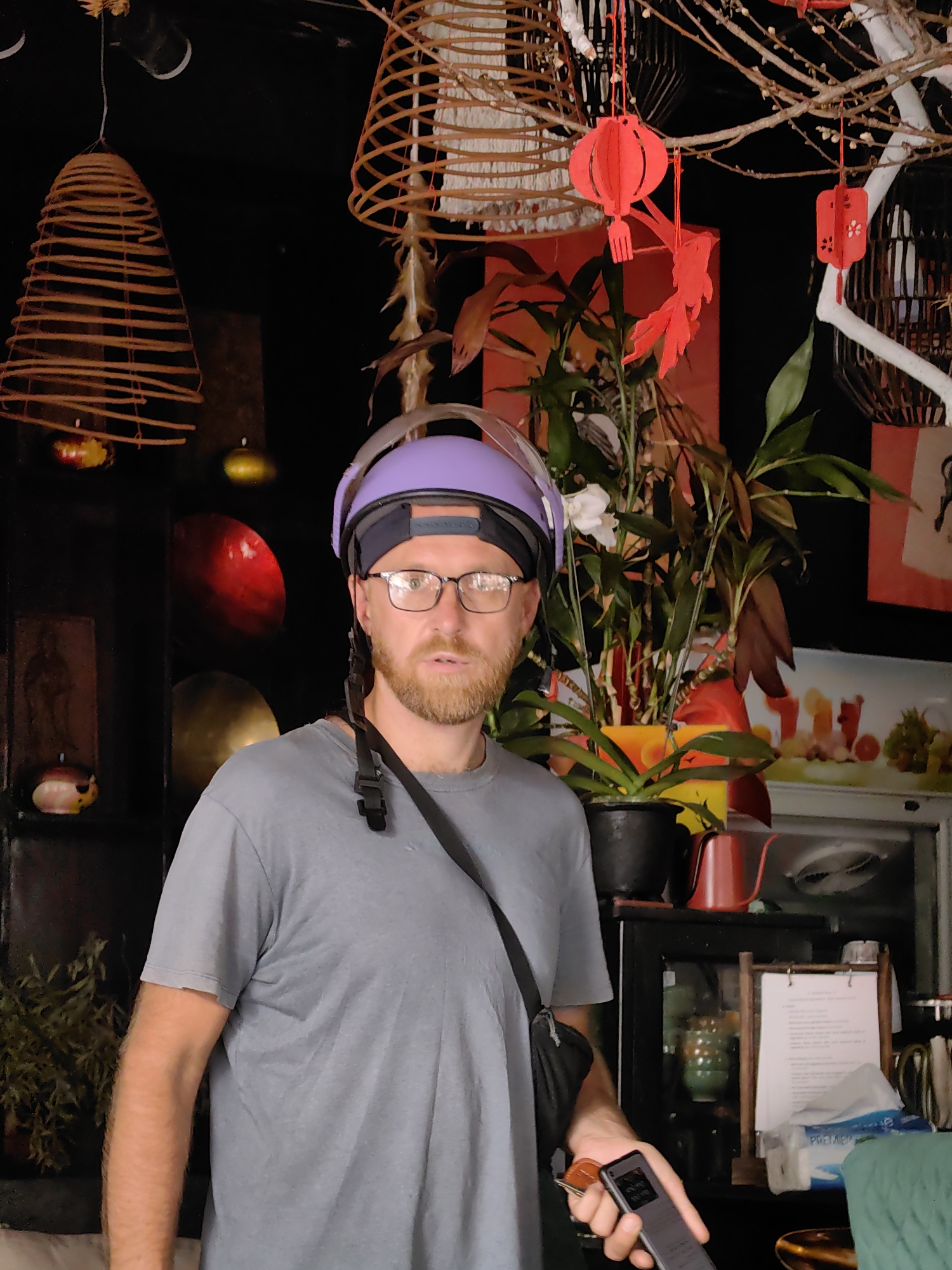 A man stands in the foreground of an interior scene, possibly in Hong Gai Ward, Vietnam. He is wearing a gray t-shirt, glasses, and a purple helmet with a black headband. A black strap of a bag or camera is slung across his body. He is holding a smartphone in his right hand.
Behind the man are various decorative elements, including woven spiral decorations hanging from the ceiling, one resembling an incense coil. There are plants with red and green foliage, and red paper lanterns are suspended from branches. A glass-front refrigerator is visible in the background, with a menu or sign on a stand in front of it. Other objects, like a red kettle, are placed on top of the refrigerator. The overall lighting is dim, creating a slightly shadowed atmosphere.