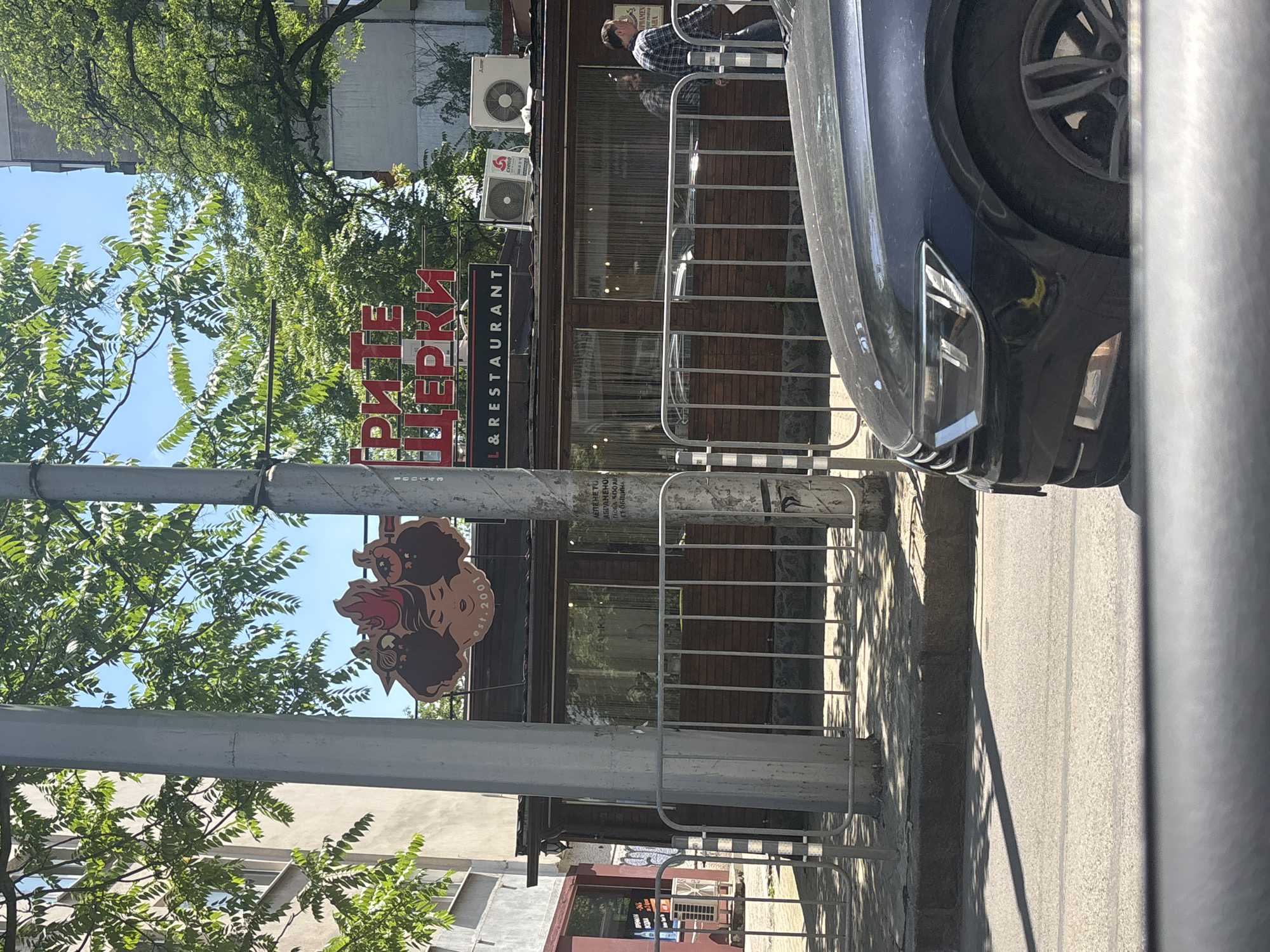 The image shows a street scene in Sofia, Bulgaria, on a sunny day. In the foreground, the front corner of a dark-colored car is visible. Behind the car, a metal fence runs along the sidewalk. Beyond the fence, a building with a wooden facade houses a restaurant. A prominent sign above the restaurant reads "ГРИТЕ ШЕРКИ" (GRITE SHERKI), with "EST. 2001" and "& RESTAURANT" visible below. Two air conditioning units are mounted on the roof of the building. Trees with green foliage are abundant, casting shadows on the ground. People are seen walking in the background near the restaurant entrance. The architecture suggests an urban environment.