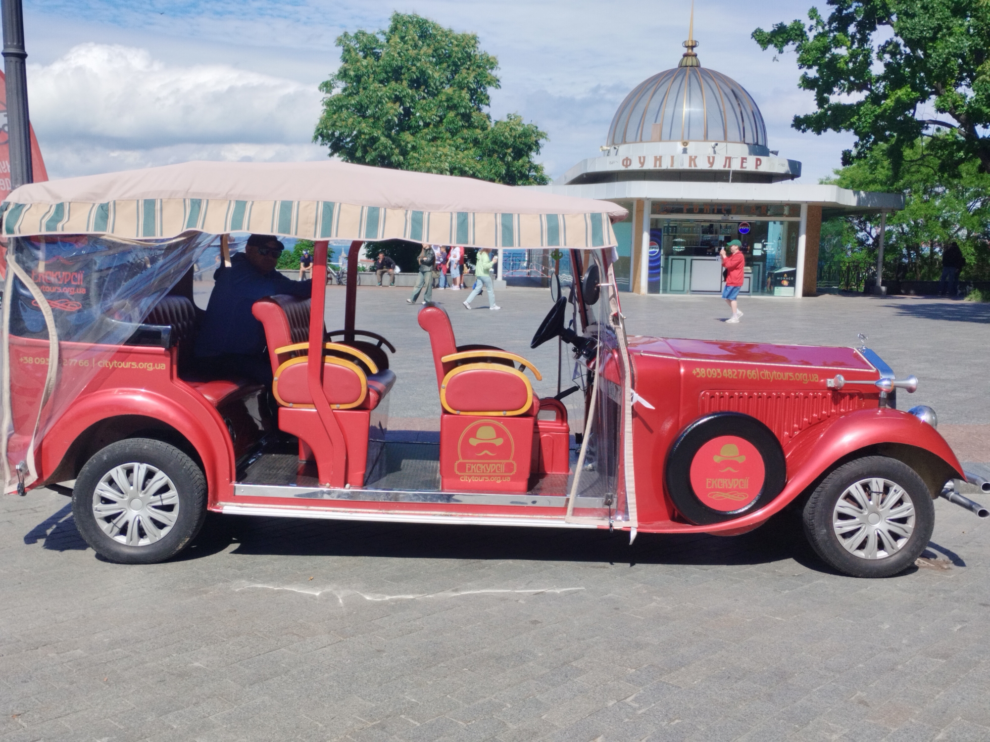 The image shows a red tourist vehicle resembling a vintage car, with a striped awning and plastic side curtains, parked on a paved surface. A driver is seated in the front, and the vehicle is labeled with "ЕКСКУРСІЇ" (Excursions in Ukrainian) and the website "citytours.org.ua" along with a phone number.
In the background, pedestrians walk near a building with a distinctive domed roof, identified as "ФУНІКУЛЕР" (Funicular in Ukrainian). Trees and a cloudy sky complete the background.
The scene is likely situated in Odesa, Ukraine, given the Ukrainian signage and the architectural style of the funicular building.