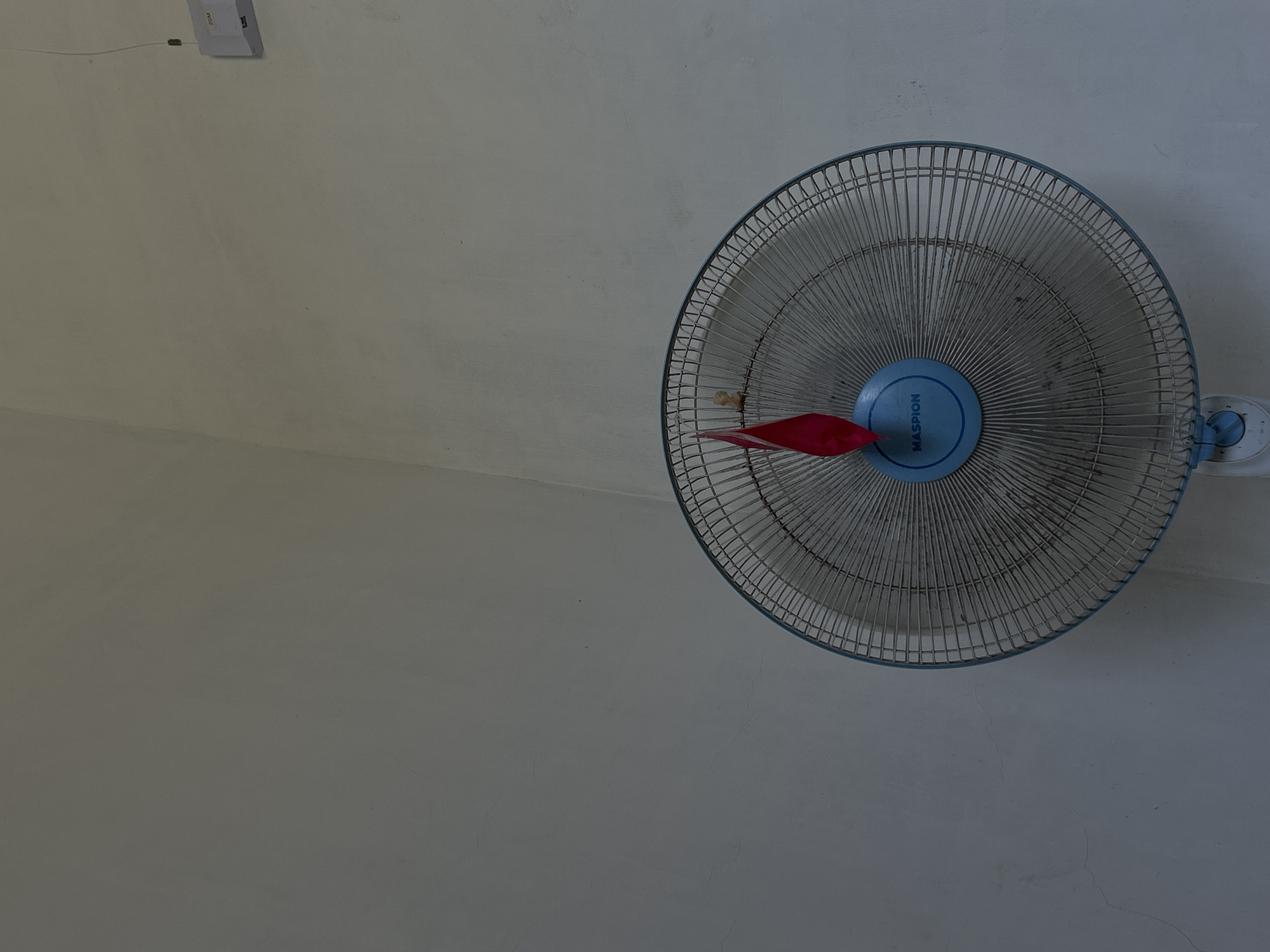 The image depicts a ceiling fan in a room. The fan is blue and silver with three red blades. The wall is white and shows signs of age and wear. A white switch box is visible in the upper left corner. The image lacks information about the location in Lhokseumawe, Indonesia, people, actions, or interactions.