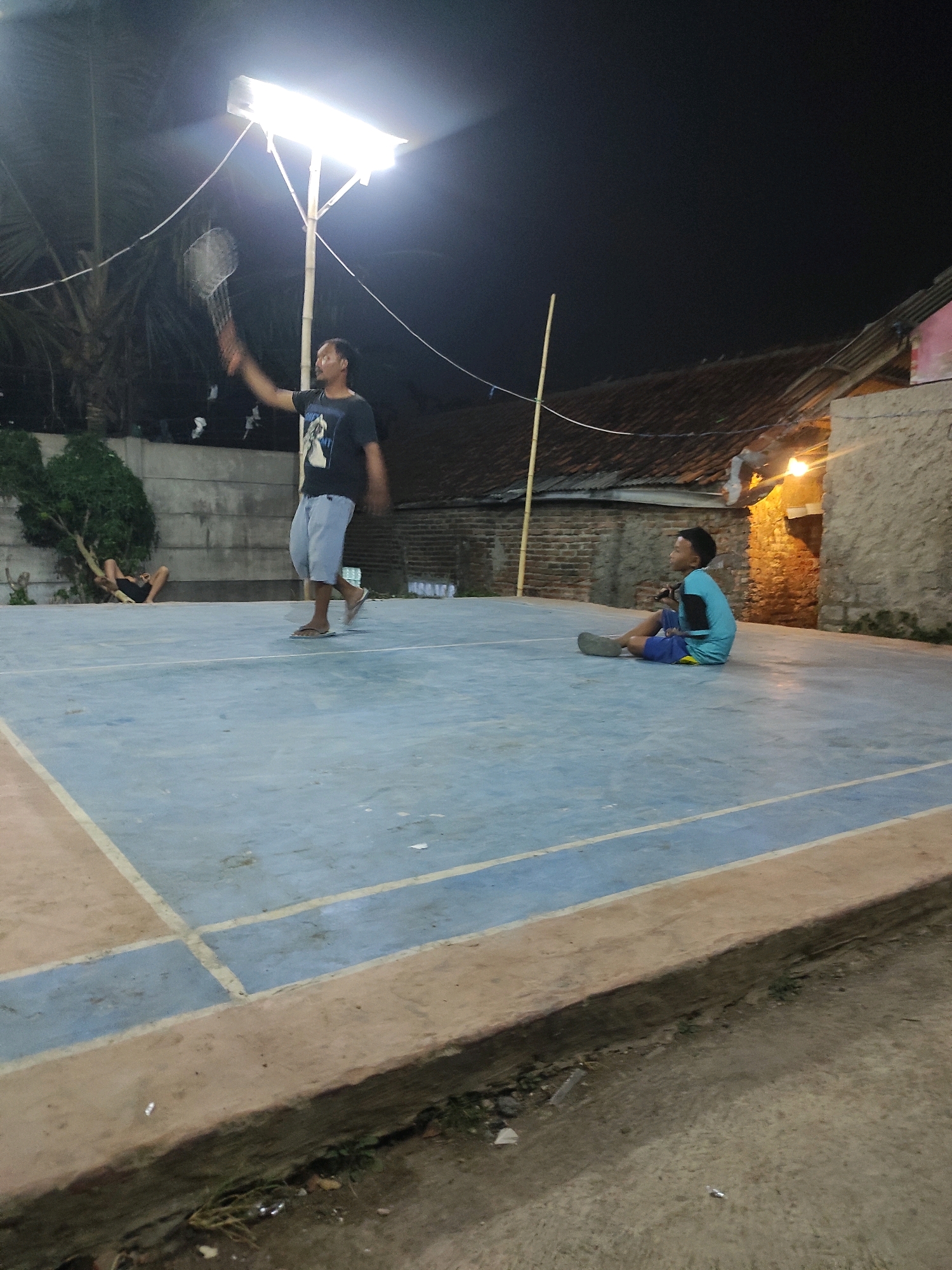 In Padalarang, Indonesia, at night, two young males are on a badminton court. One male, wearing a black t-shirt with a graphic and light-colored shorts, is in the process of swinging a badminton racket, illuminated by a bright overhead light. The other male, dressed in a blue t-shirt and dark shorts, is seated on the court. In the background, there is a palm tree, a concrete wall, and a residential building with a tiled roof, also lit by outdoor lighting. The court surface is predominantly blue with tan borders.