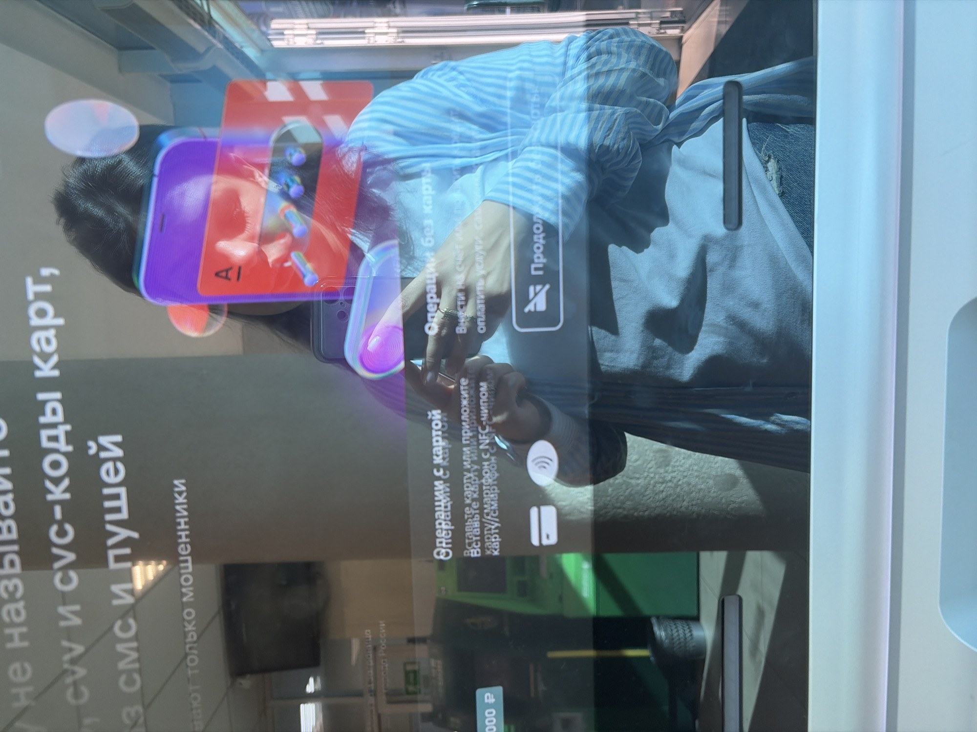 An individual is reflected in the screen of a self-service terminal or ATM, located in Sochi, Russia. The reflection shows a person, likely female, with dark hair, wearing a light blue and white striped shirt over a lighter undershirt, with visible denim fabric, possibly jeans, featuring a distressed area. Her face is partially obscured by superimposed digital graphics, including a purple smartphone icon, a red bank card icon with an "A" symbol, and luminous circular elements. She holds a dark-colored smartphone in her left hand (viewer's right).
The screen displays Russian text, including a fraud warning at the top: "Don't reveal CVV and CVC card codes, SMS and push notifications... only fraudsters ask for them." Lower sections offer "Card operations" ("Insert card or attach card/smartphone with NFC chip," indicated by card and NFC icons) and "Cardless operations" ("Deposit to account or top up... pay for services, communications..."). A "Continue" button with a disabled icon is also visible.
In the background reflection to the left, a green ATM or self-service terminal is visible, displaying "1 000 ₽" (1,000 Russian Rubles) on its screen. The environment suggests an indoor public space, possibly a bank, with a light-colored ceiling, linear lighting, and a partially visible wall and floor.