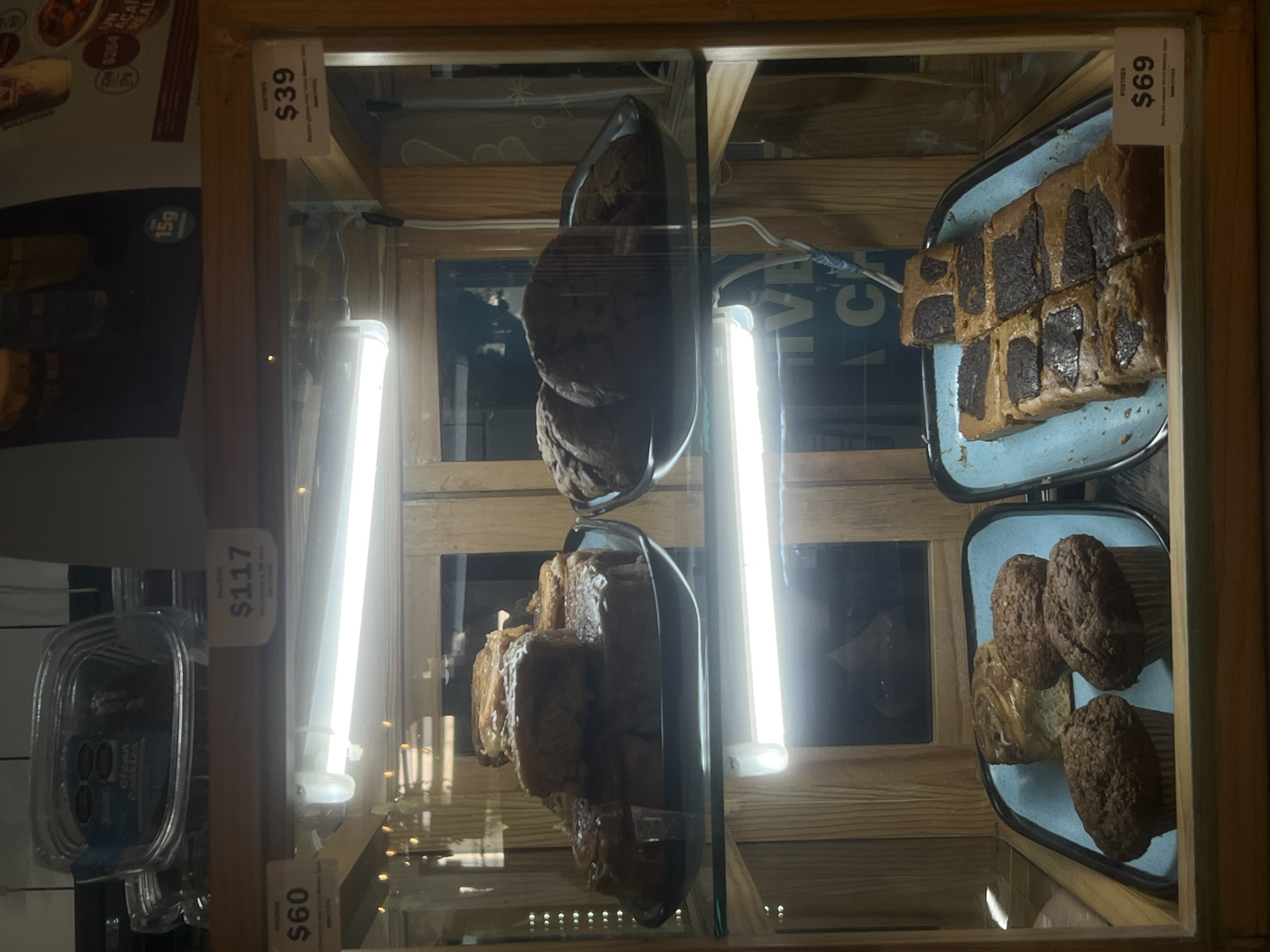 This is an image of a display case filled with baked goods, possibly in a cafe or bakery. There are three types of items visible: large cookies stacked on a tray, pastries (likely brownies) cut into squares on a blue tray, and muffins on a blue tray. Price tags are visible next to each item, indicating their cost. One tag reads "$39", another reads "$69", and another reads "$60", and "$117". The display case is made of wood and glass. The lighting in the case is provided by fluorescent tube lights, which cast a bright glow on the pastries. The photo may have been taken in Mexico City, Mexico, based on the OCR text in the background.