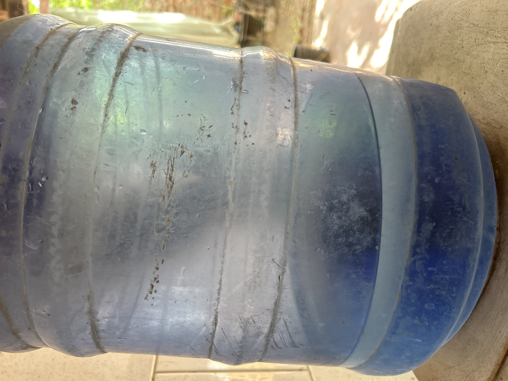 A close-up, low-angle shot shows the lower half of a large, transparent blue plastic water jug. The jug has prominent horizontal ridges and appears dusty and somewhat dirty. The water inside is visible, filling the bottom third of the jug, with a darker blue hue at the base and a lighter, clearer blue towards the top. The jug is resting on a textured, light-colored wooden surface. In the background, a lightly-tiled wall is visible to the left, and a blurry outdoor scene with greenery and a hint of brick structure to the right suggests it is located outdoors in Santa Ana, Venezuela. Sunlight is casting dappled shadows across the scene.