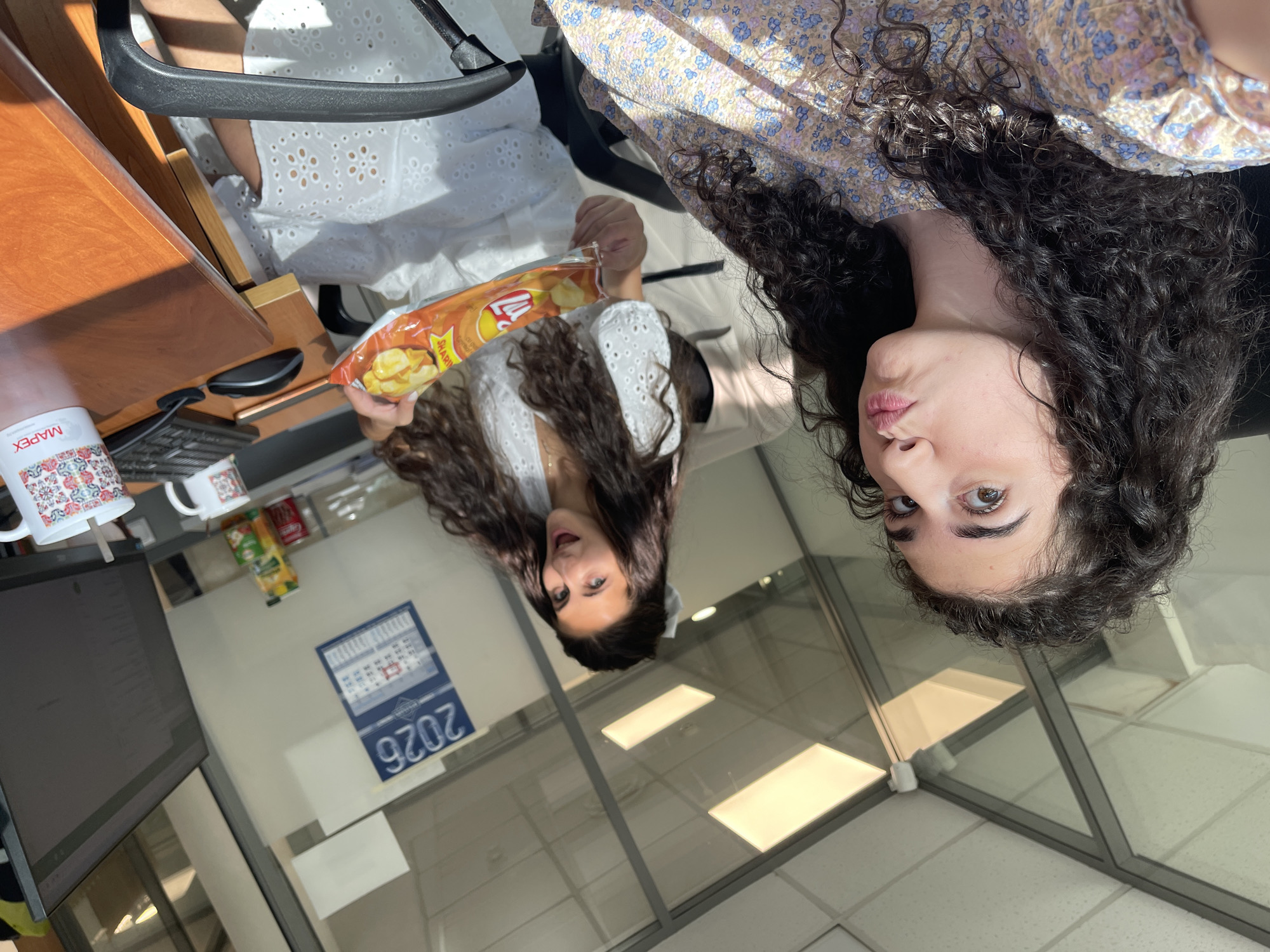 Two young women are in an office environment, likely in Sofia, Bulgaria, as indicated by a calendar on the wall displaying "2026" and the branding on a mug that says "MAPEX." The woman in the foreground, on the left, has dark, curly hair and is wearing a floral print dress. She is making a "pucker" face towards the camera, holding the camera herself in a selfie-like pose. The woman in the background, on the right, also has long, dark hair and is wearing a white eyelet dress. She is holding a bag of chips and appears to be offering them to the camera or the other woman. The office has glass partitions, fluorescent lighting, and desks with computer equipment. There are mugs, a keyboard, a mouse, and a juice box visible on the desk. The overall impression is a casual, possibly break-time moment within a professional setting.