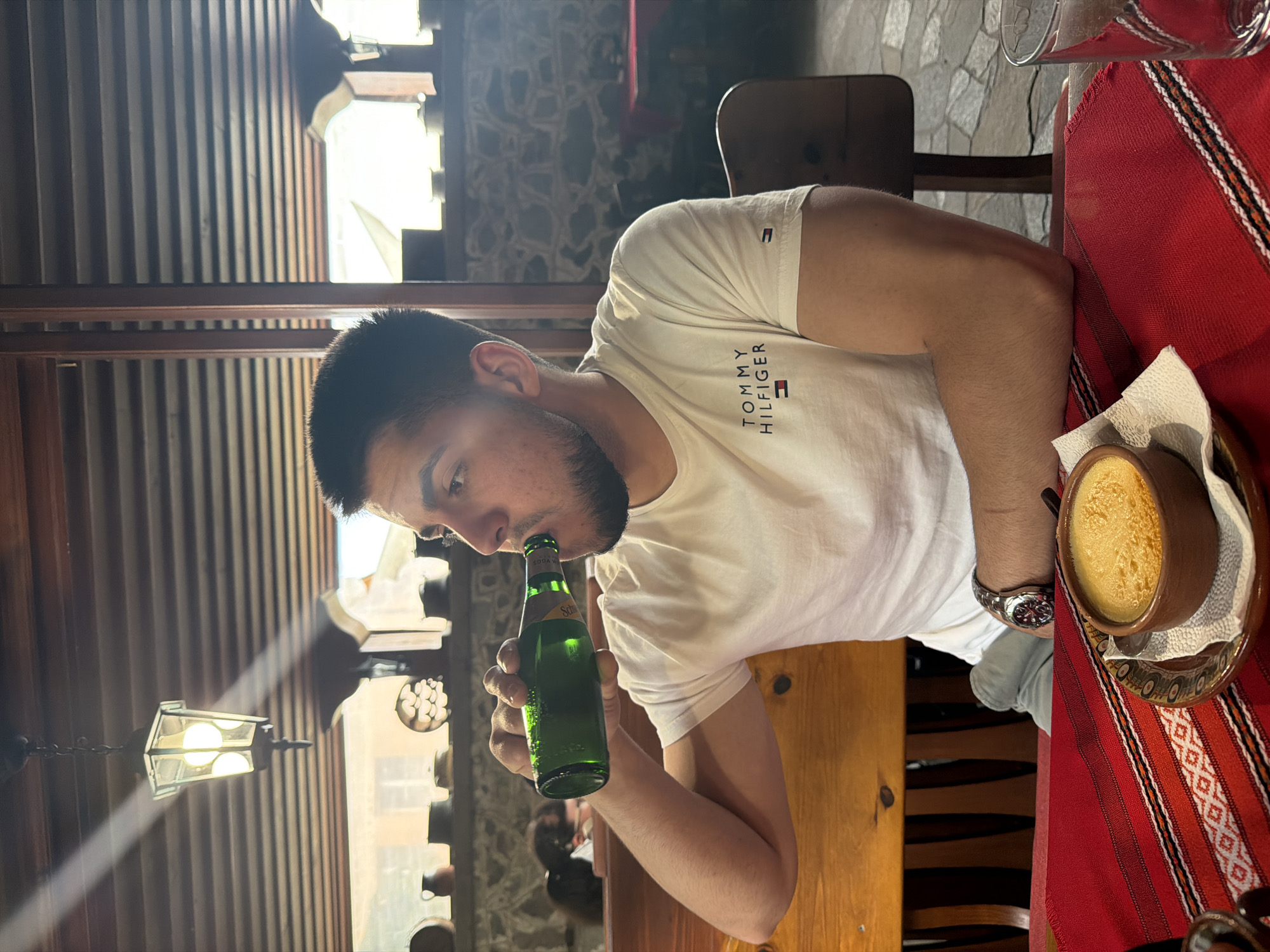 A medium eye-level indoor shot depicts a man drinking a green glass bottle of soda in a restaurant in the city of Panagyurishte, Bulgaria. The man has dark hair and a beard and is wearing a white Tommy Hilfiger T-shirt and a watch on his left wrist. He is seated at a wooden table with a red tablecloth featuring a white and black stripe pattern. A ceramic bowl containing food sits on the table. The background features stone walls, windows with wooden shutters, and a decorative light fixture.