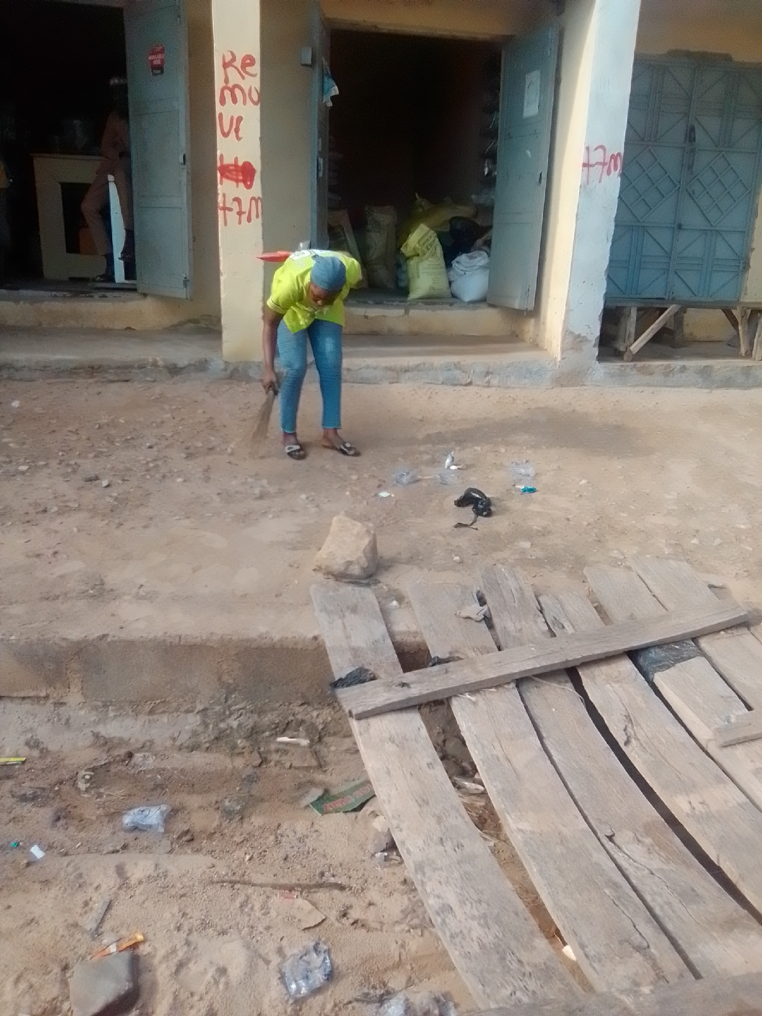 A person wearing a yellow shirt and blue jeans is sweeping the ground in front of a building. The building appears to be a shop with an open doorway and a closed door. There is writing on the wall of the building that says "Re mo ve +7m". The ground is made of dirt and there is some debris scattered around, including plastic bottles and a rock. A wooden structure is visible in the foreground. The scene is outdoors, and the lighting suggests it is daytime. The location is identified as Galadima, Nigeria.