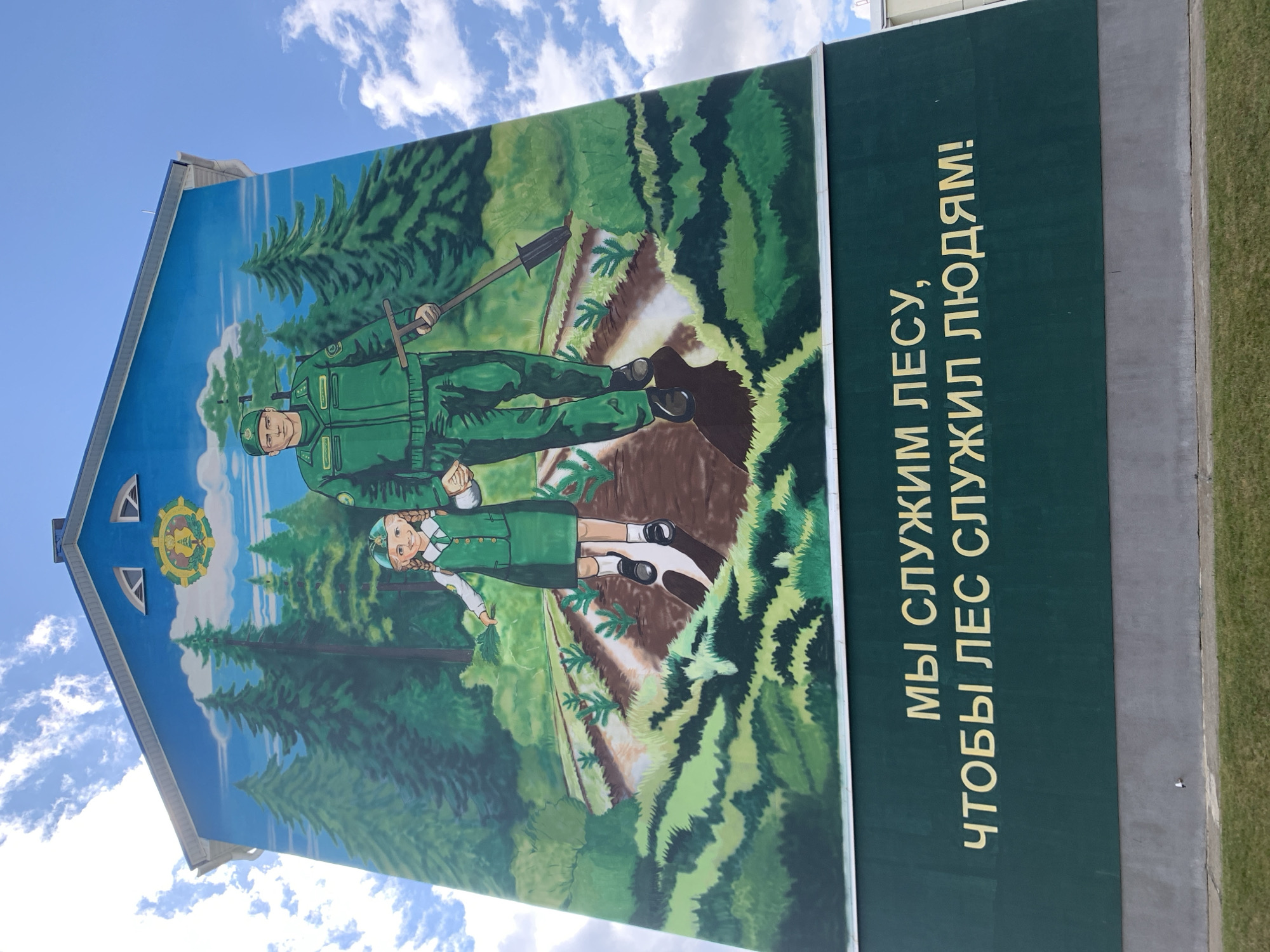 This is a mural painted on the side of a building in the city of Kapyĺ, Belarus. The mural depicts a man and a young girl in green uniforms, standing in a forest setting with young saplings planted in rows in front of them. The man is holding a shovel, and the girl is holding a small branch. Above them, a circular emblem with a green tree and a golden wreath is painted on the blue sky. The bottom portion of the mural features green text in Cyrillic, which translates to "We serve the forest so that the forest serves people!" The building itself is light grey at the base and has a white roofline. The sky is blue with white clouds.