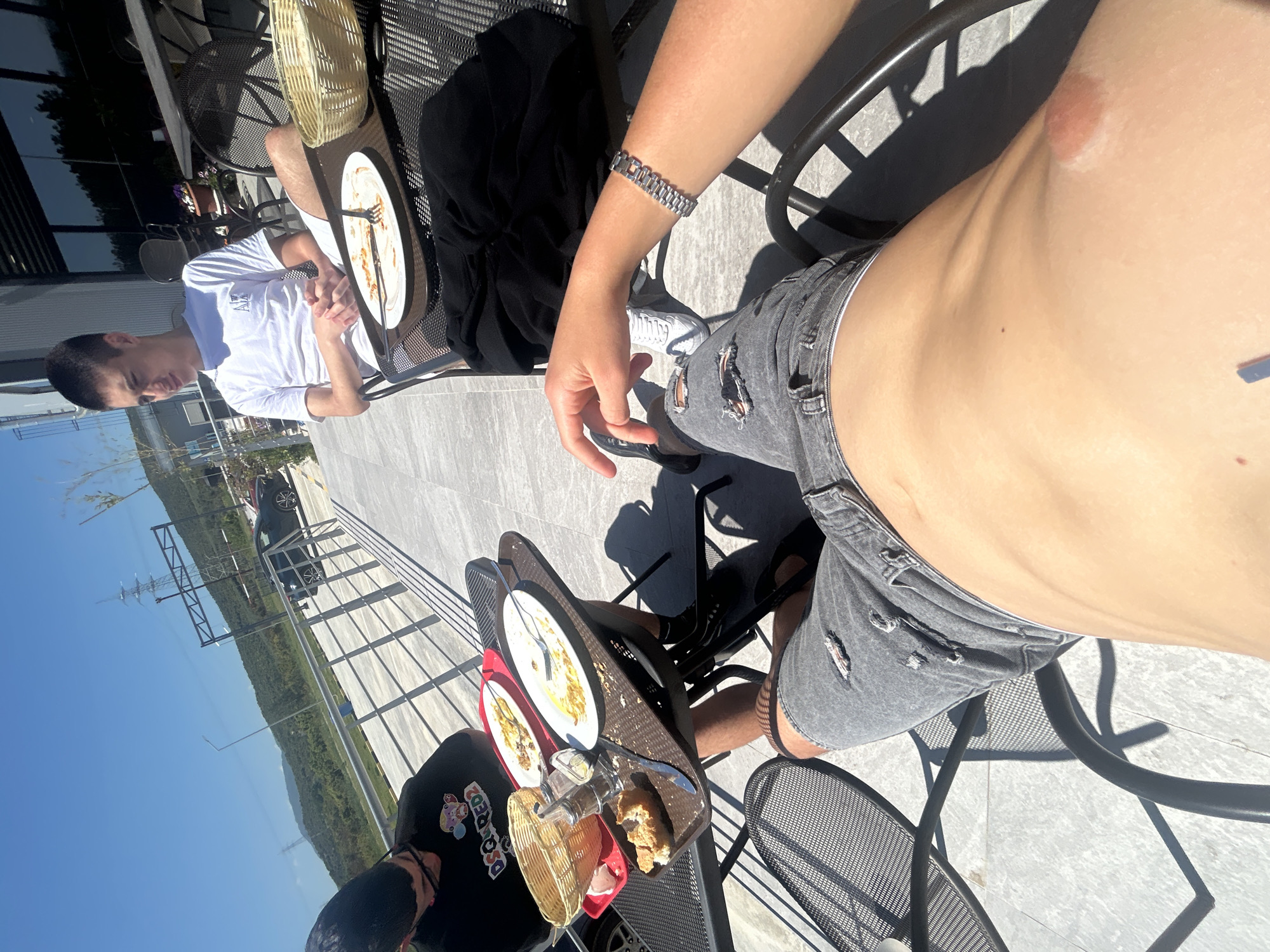 Captured in Νέα Κερδύλια, Greece, the image presents a top-down view of an outdoor dining setting. Two individuals are visible: one wearing a white shirt and the other in ripped grey jeans, a silver bracelet, and a black top with cartoon characters on it. A tray holds dishes with remnants of a meal, utensils, and a basket. Metal chairs surround the table. In the background, a sunny sky, green vegetation, power lines, a road, and parked vehicles are discernible.