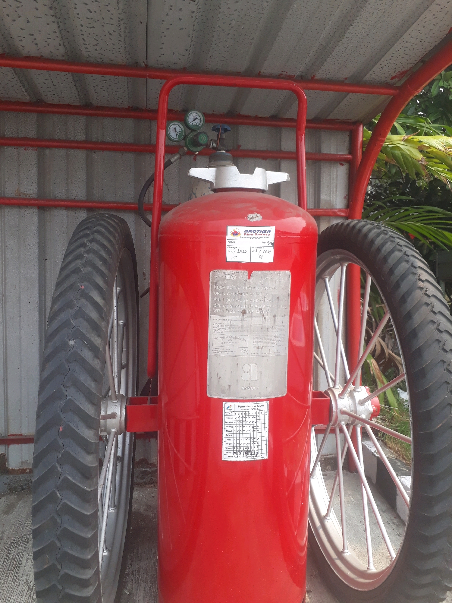 The image shows a bright red fire extinguisher mounted on a cart with two large spoked wheels. The fire extinguisher has a silver top and a silver label with white text. The cart's frame is also red, matching the extinguisher. Above the extinguisher, there are three pressure gauges. The scene appears to be outdoors, with a corrugated metal roof overhead and some greenery in the background. The ground is concrete. This fire extinguisher is likely located at a facility in the city of Pangkalpinang, Indonesia, given the context provided.