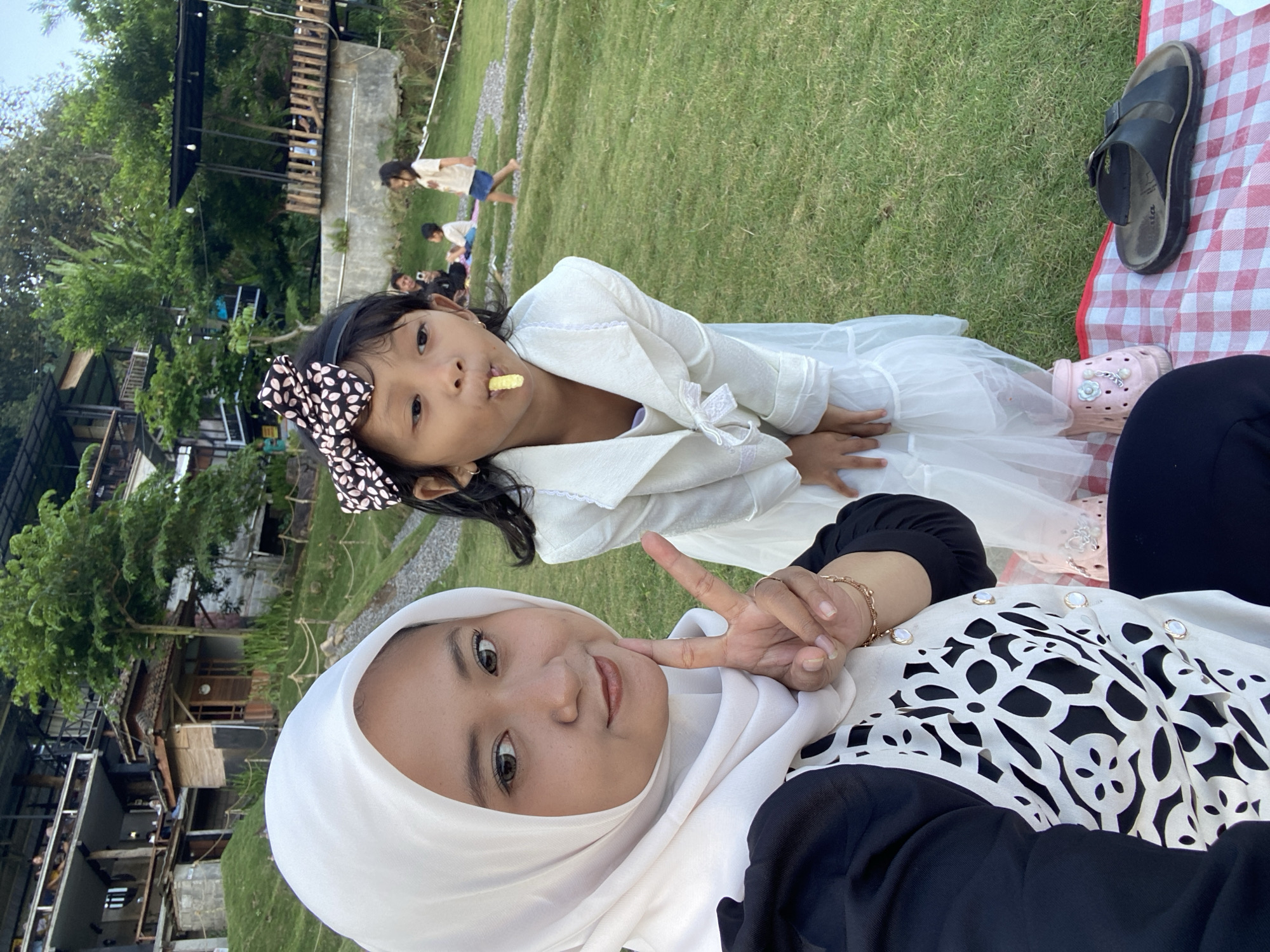 In a grassy outdoor area with multi-level structures in the background, a woman wearing a white hijab and a black top with a white patterned vest is taking a selfie. Beside her, a young girl in a white dress and jacket with a black and white polka dot bow in her hair is leaning forward, holding a light-colored snack in her mouth. The woman is making a peace sign with her right hand. In the background, several other people, including children, are visible on the grass and on elevated platforms. The environment suggests a park or recreational area.