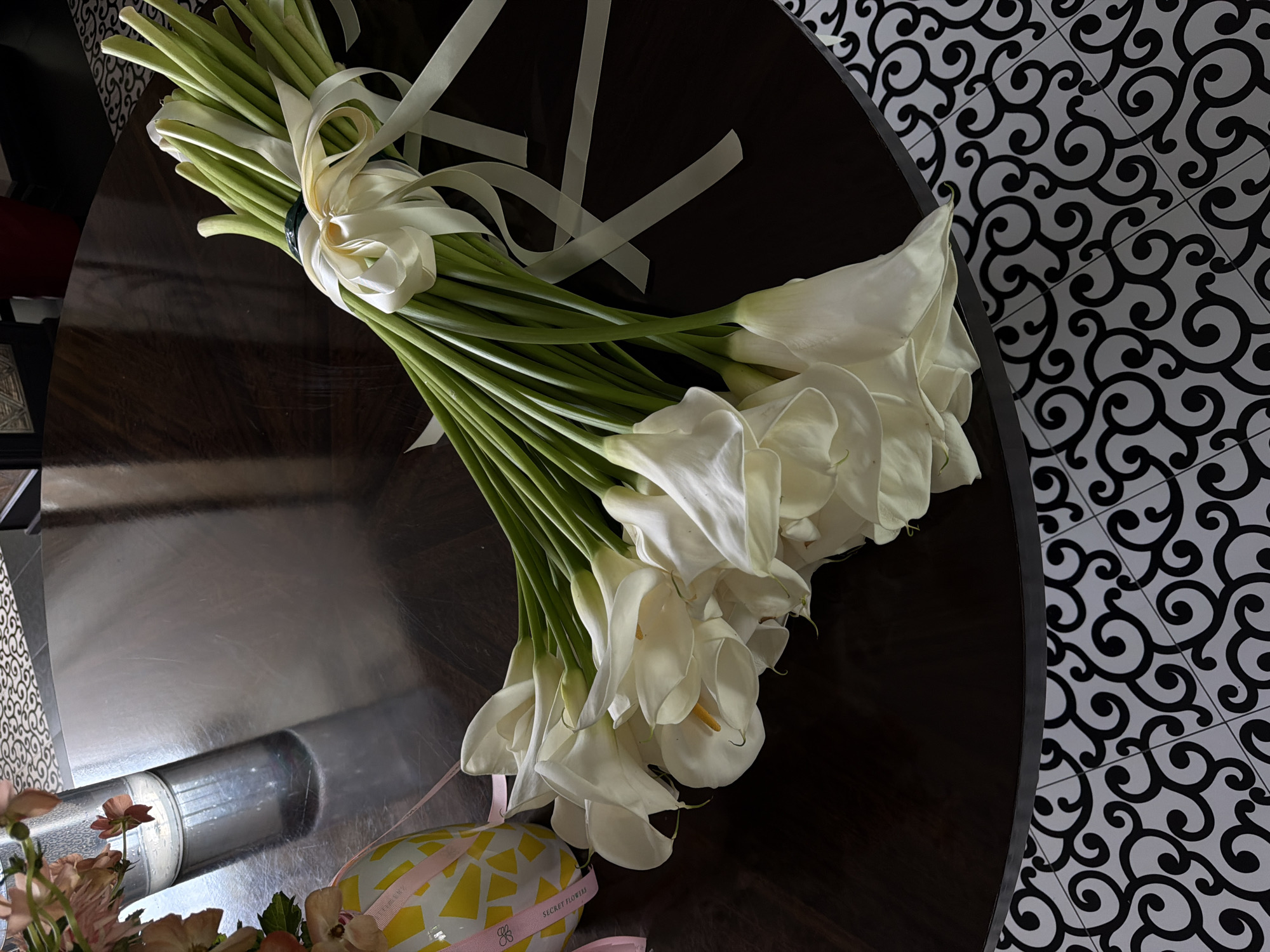 A bouquet of white calla lilies tied with cream-colored ribbons lies on a dark, circular wooden table. The table has a reflective surface and is placed on a floor with black and white ornate tilework. To the left of the bouquet, a glass vase with a few pink flowers is visible. A decorative yellow and white cylindrical object with pink ribbon detailing is also present on the table. The background is dark and out of focus. The scene appears to be indoors, potentially in a home or a flower shop. There are no people in the image. The location is not specified in Bishkek City, Kyrgyzstan.