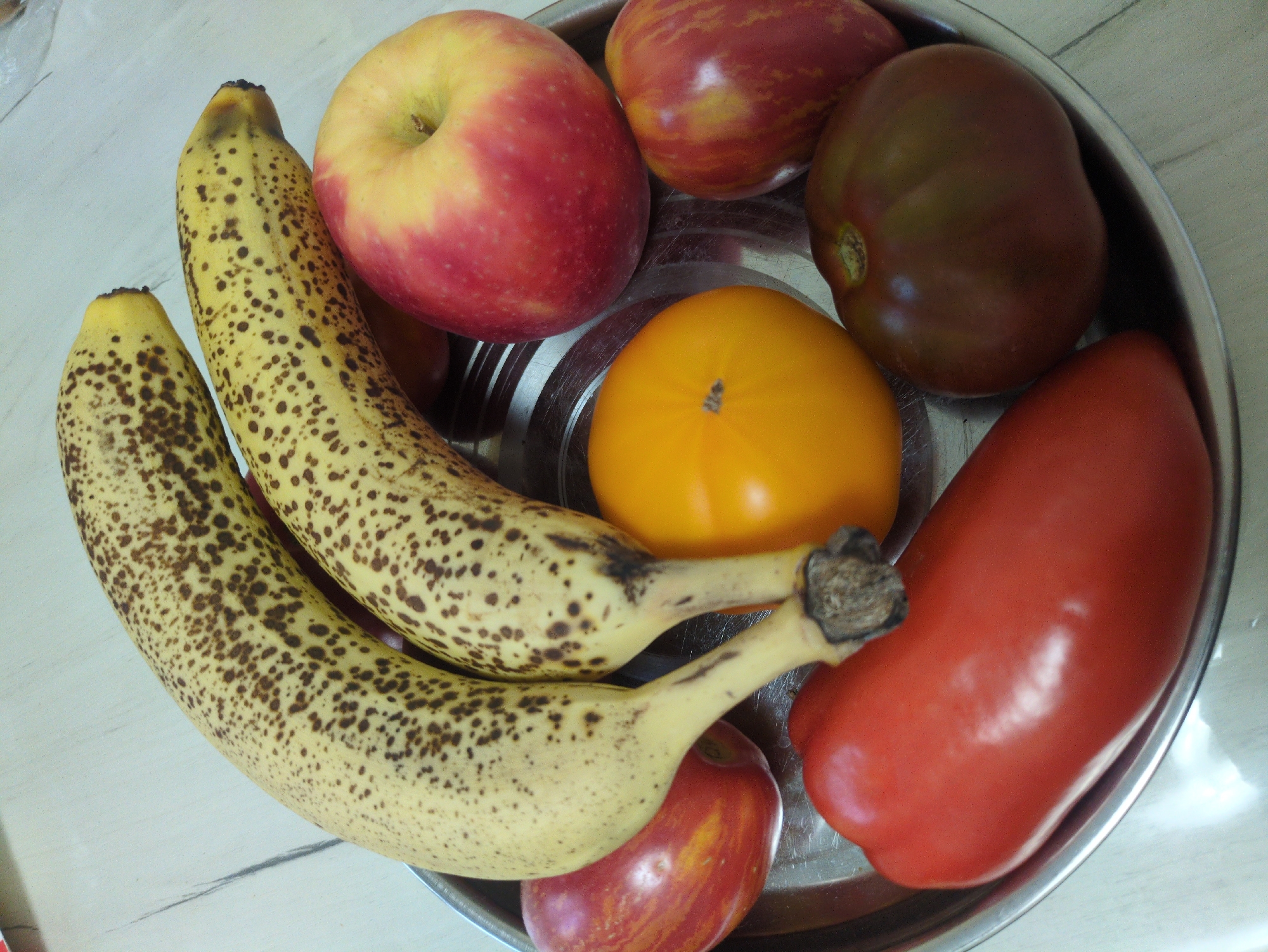 A metal bowl filled with a variety of fruits and vegetables sits on a light-colored, textured surface. The items in the bowl include two ripe bananas with brown spots, two red-striped apples, a bright orange tomato, a red bell pepper, and a dark-colored tomato. The bananas are arranged diagonally across the top of the bowl, with the red-striped apples positioned to the left and right of the bottom. The orange tomato is in the center, slightly behind the red bell pepper, which occupies the bottom left of the bowl. The dark-colored tomato is in the bottom right. The lighting suggests an indoor setting. There are no people visible in the image, and no specific actions or interactions are depicted. The scene is static. The image was taken in Kulebaki, Russia.