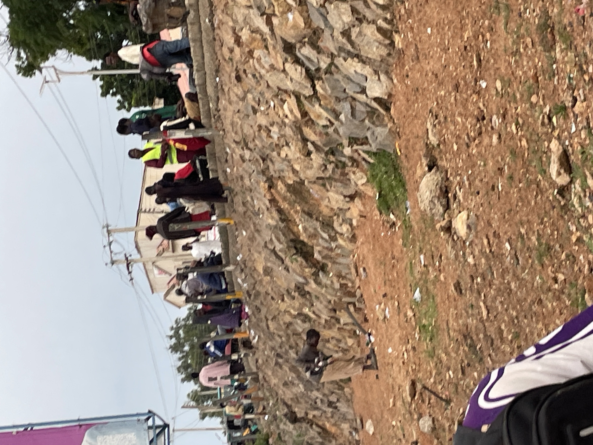 The image depicts a scene in the city of Jos, Nigeria, characterized by a rocky incline bordering a dirt path. Several individuals are present in the frame. In the foreground, a person in beige pants and a dark top bends over, seemingly engaged with an object on the ground. Further up the incline, a group of people are gathered. Some are seated on concrete steps, while others stand. Among them, a person wearing a bright yellow vest stands out. Buildings and trees are visible in the background, suggesting an urban or semi-urban environment. The ground is covered with dry earth and scattered debris. The overall atmosphere appears to be one of casual congregation.