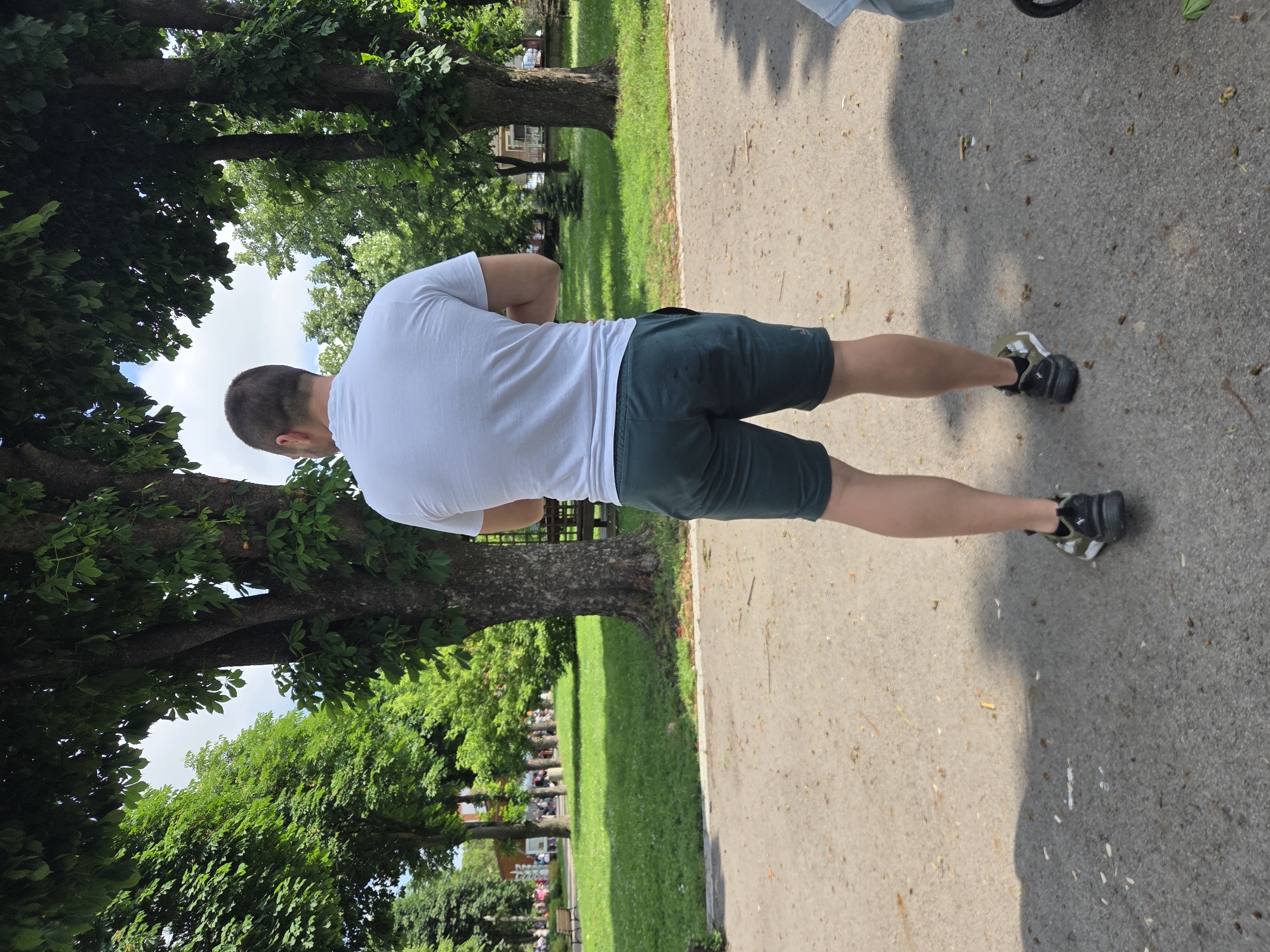An adult male is observed from the rear, standing on a paved path within a park in Elin Pelin, Bulgaria. He is dressed in a white short-sleeved t-shirt, dark green athletic shorts, and black and olive green sneakers. His arms are positioned behind his back. The grey path is scattered with leaf litter and small organic debris. A partial view of a light blue item and a wheel, likely from a stroller, is visible to the man's immediate right. The scene's midground features a manicured green lawn and numerous large trees with dense foliage, indicating a summer environment. Sunlight projects shadows onto the path. In the background, additional trees and architectural structures are visible, consistent with a public park situated within a town or city. Distant figures are faintly discernible on the far left. The sky, seen through the tree canopy, suggests a bright day with minimal cloud cover.
