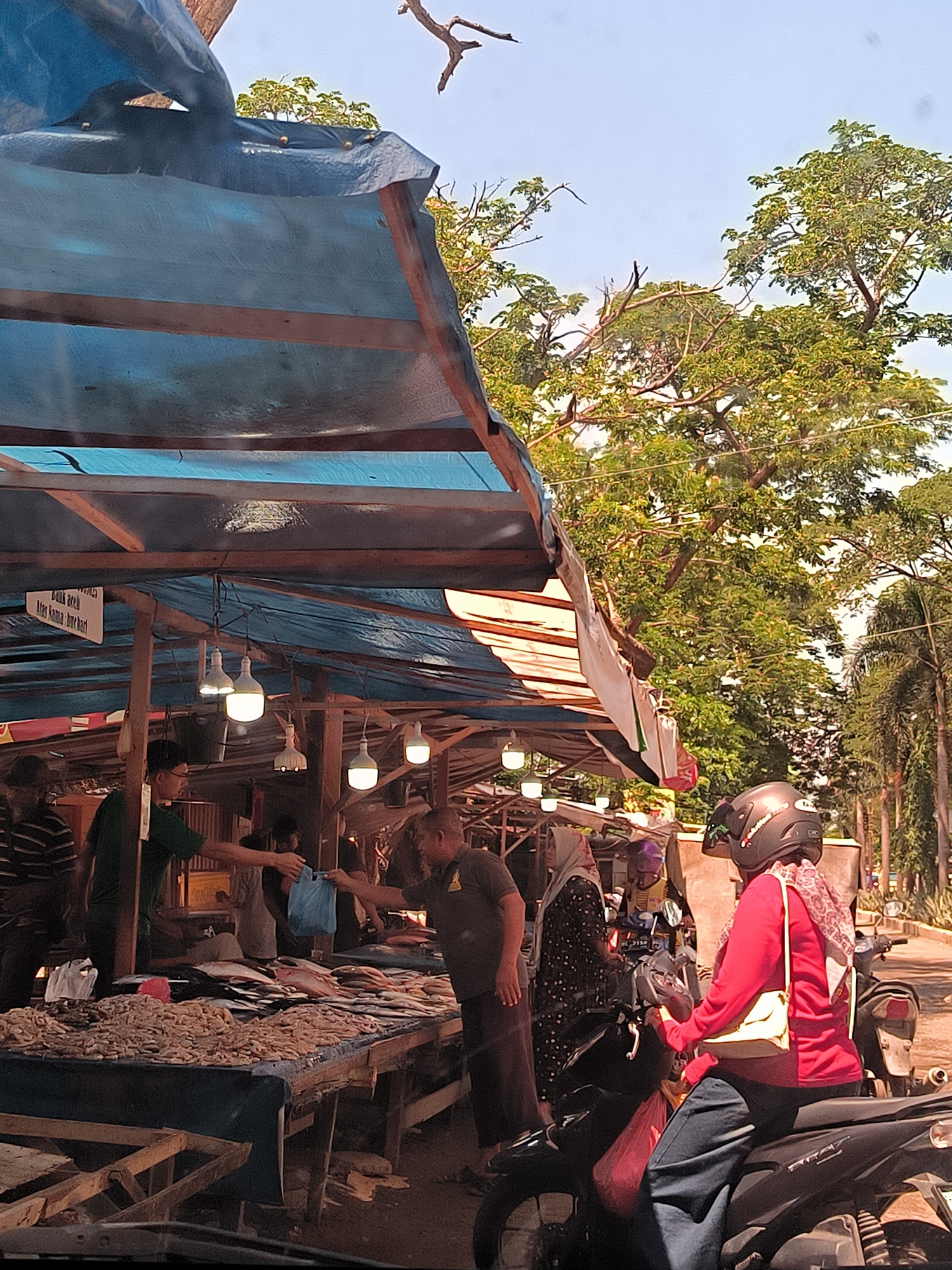 The image depicts an outdoor market scene in Lhokseumawe, Indonesia. The market is covered by a blue tarp suspended on wooden beams, with several bare light bulbs illuminating the stalls. Tables laden with dried fish and what appear to be small shrimp are visible. People are engaged in transactions, with a man in a green shirt handing a blue plastic bag to another man across a table. A woman wearing a red jacket and a helmet is seated on a black motorcycle in the foreground, facing away from the camera, with a light-colored purse slung over her shoulder. In the background, lush green trees and palm trees line a paved road, suggesting an urban or semi-urban environment. The sky is bright and clear, indicating daylight.