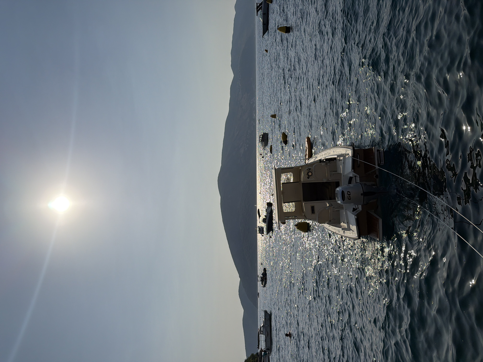 This image shows a view of the sea in Zelenika, Montenegro, with the sun shining brightly in the sky. In the foreground, a small white motorboat with a blue hull is moored. It has an outboard motor with the number "60" visible on it. In the background, several other boats are scattered across the water, and a chain of yellow buoys stretches across the bay. Beyond the water, a range of dark, tree-covered hills form the horizon. The water is choppy and reflects the sunlight, creating a shimmering effect. The sky is a clear, pale blue, with the sun appearing as a bright, slightly blurred orb. A person can be seen swimming in the water to the left of the frame. The scene appears to be a serene coastal landscape during daytime.
