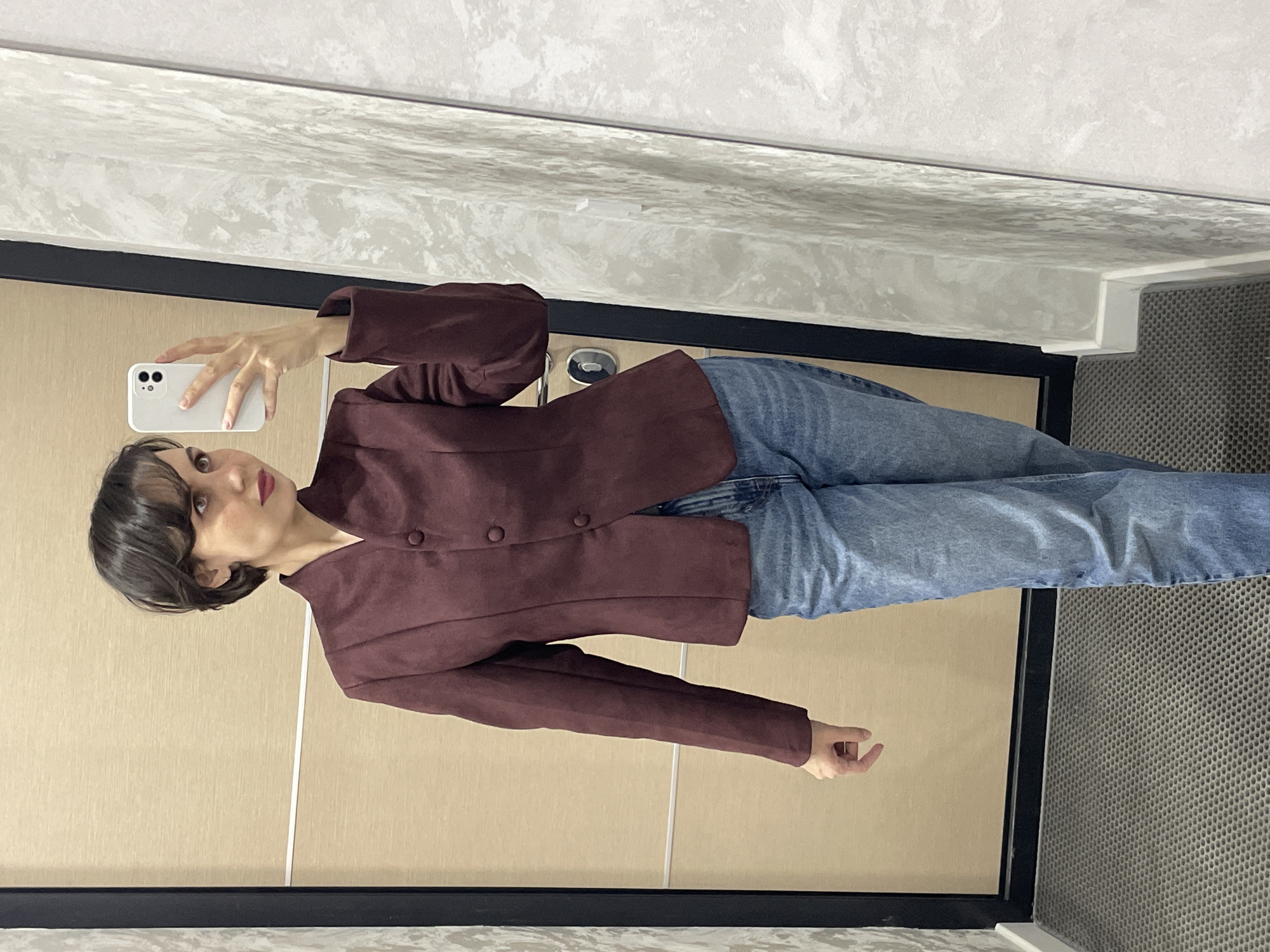 A person stands in what appears to be a bathroom in Izhevsk, Russia, based on the design and materials often used in the city. They are taking a selfie using a white iPhone. The person is wearing a burgundy-colored jacket with multiple buttons and light blue jeans. The walls are a light beige, and the ceiling has a marbled texture. The floor appears to be covered with a gray, textured material.