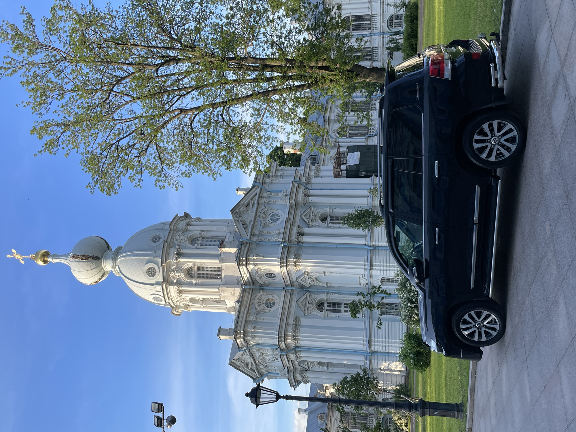 The image depicts the Smolny Cathedral and Convent complex, located in St. Petersburg, Russia, under a clear blue sky. The prominent subject is the Smolny Cathedral, a large, multi-domed Baroque structure predominantly painted light blue with white architectural details, including ornate columns, pilasters, and decorative window surrounds. Its main dome culminates in an onion-shaped cupola, topped with a golden sphere and a golden Orthodox cross. To the left of the main cathedral, an adjacent building within the complex shows scaffolding and construction barriers on its exterior.
In the foreground, on a grey paved surface composed of square tiles, a dark-colored (likely black) Toyota Land Cruiser 200 series SUV is parked, facing slightly left. The vehicle features chrome side steps, roof rails, tinted windows, custom multi-spoke wheels, and a rear bumper guard. A "Executive Lounge" badge is visible on the rear tailgate.
The area surrounding the cathedral includes a well-maintained green lawn. A large deciduous tree with light green foliage and multiple trunks stands to the right of the cathedral, partially obscuring other buildings in the complex. Smaller trees and bushes are scattered throughout the grassy areas. A black, ornate lamppost with a single light fixture is positioned on the left side of the image, between the parked car and the cathedral. High on the left side, against the blue sky, two black security cameras are mounted on a pole. The scene is brightly illuminated by natural daylight, with no individuals present.