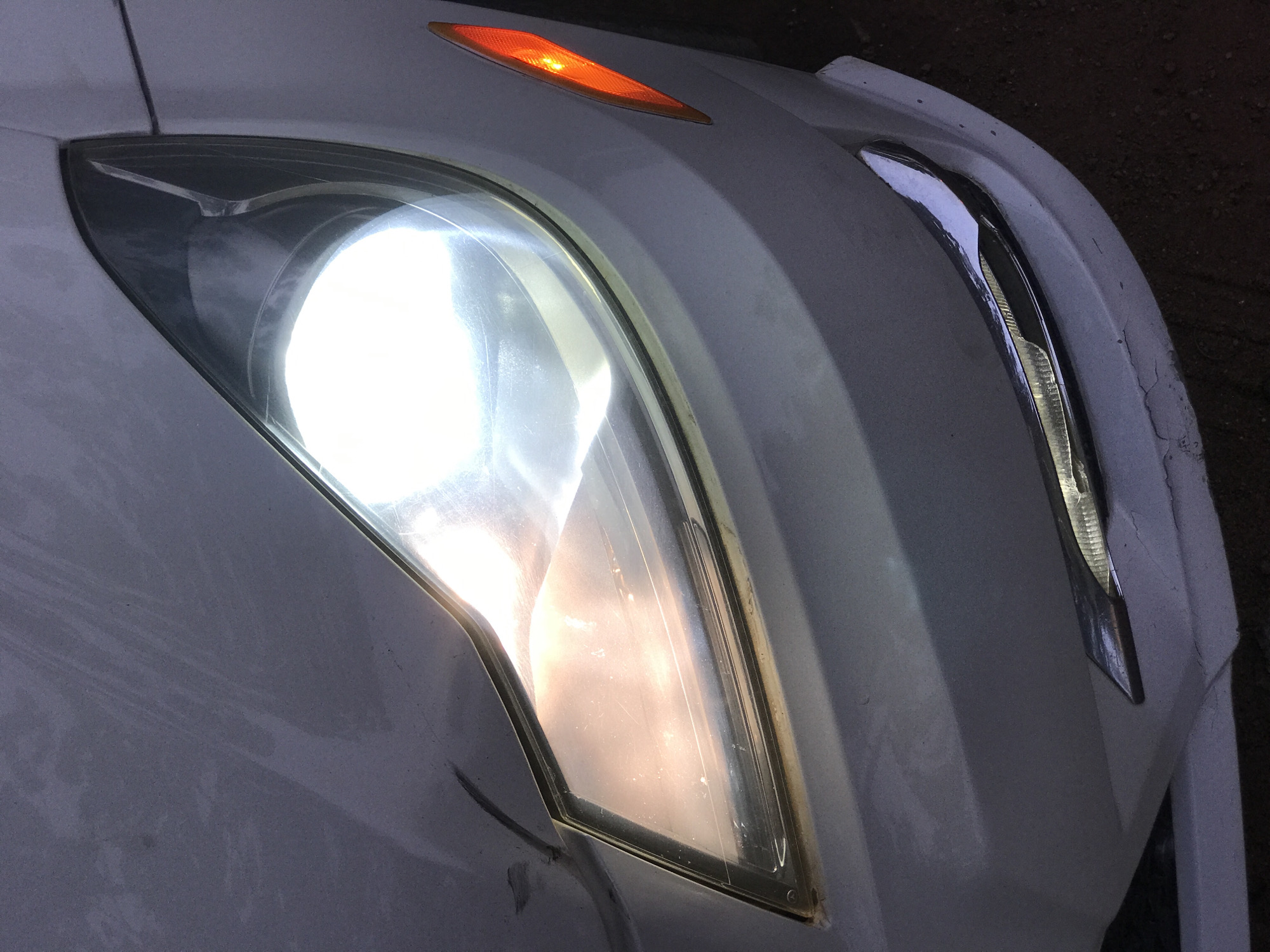 The image captures a close-up of the front section of a white car, likely at dusk or night, given the illuminated lights and dark background. The car's left headlight is brightly lit with a white glow, indicating it is on. Above and slightly to the left of the main headlight, a smaller, orange turn signal or marker light is also illuminated.
The white paint of the car's body and fender shows signs of wear, including scuffs, dirt, and some darker streaks. Closer inspection, as revealed by the cropped images, highlights several notable details:
- The main headlight's clear plastic cover appears somewhat hazy, scratched, and shows accumulated dirt or film.
- The adjacent car body near the headlight and especially on the right fender exhibits paint damage, including cracks and chips, suggesting wear and tear or minor impact.
- A chrome trim piece located on the far right side of the car's front also appears damaged or worn, with visible indentations and reflections indicating its metallic nature.
- The car's surface generally appears dusty or dirty.
There is no visible text in the image. The scene takes place in Galadimawa Village, Nigeria.