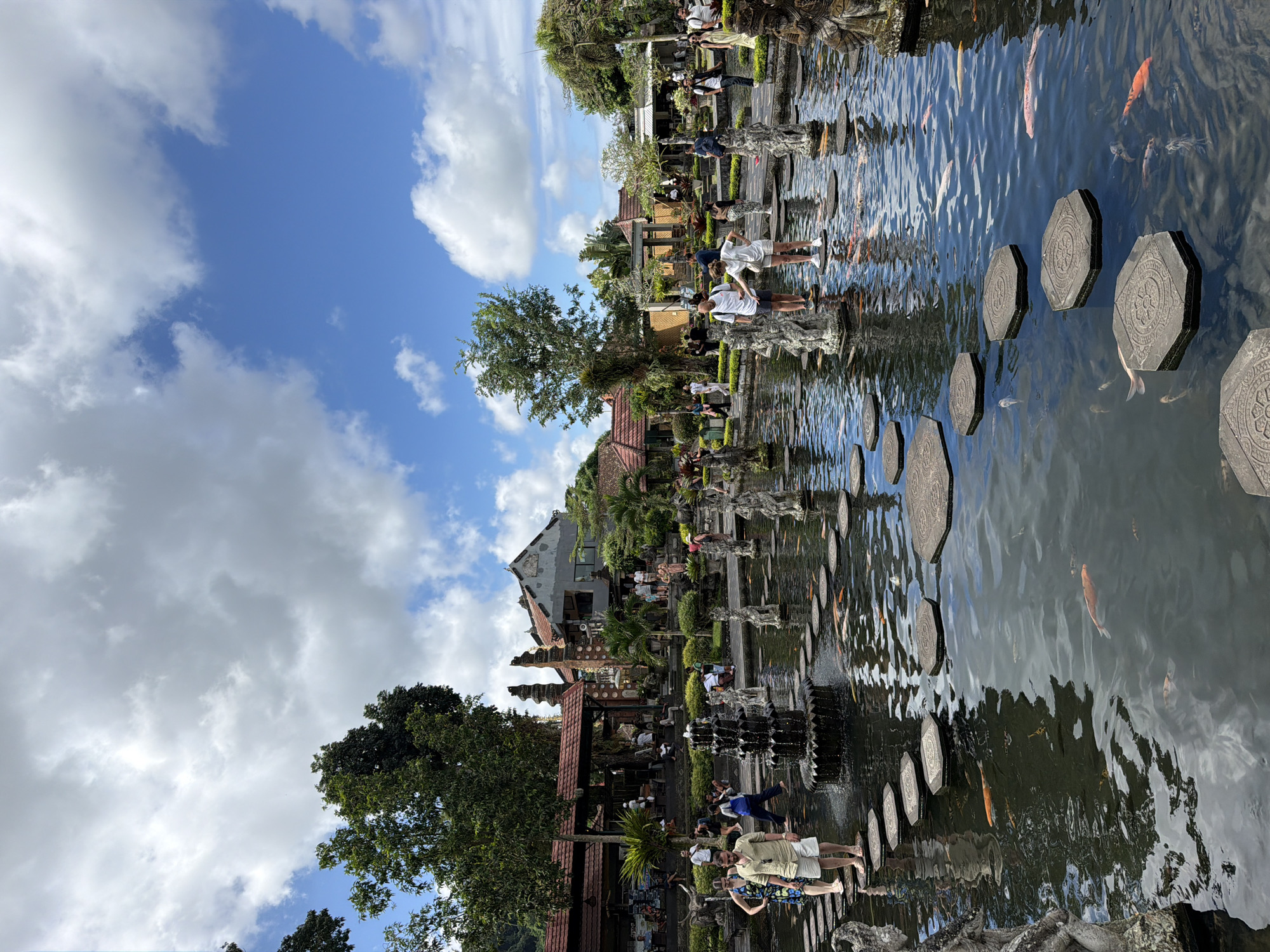 This image captures the Tirta Gangga Water Palace in Abang, Indonesia, on a partly cloudy day. The foreground features a large pond with numerous koi fish swimming in the murky water. Stepping stones, octagonal in shape with decorative carvings, are arranged in a path across the pond. Several stone statues, likely of mythical figures, are placed along the pond's edge and within the water, acting as supports for more stepping stones.
In the middle ground, a tiered black fountain with water cascading down its levels is positioned on a stepping stone. Around the pond, people are visible, some walking on the stepping stones, others observing the pond or the surrounding architecture. A man in a light-colored short-sleeved shirt and shorts is standing on a stepping stone, while a woman in a patterned dress is to his left. Further back, more individuals are seen walking along paved paths and near buildings.
The background reveals traditional Balinese architecture, including buildings with red-tiled roofs, intricately carved wooden elements, and stone towers. Lush green trees and palm trees are scattered throughout the scene, adding to the tropical ambiance. The sky is a vibrant blue with scattered white and gray clouds. The overall scene depicts a popular tourist destination with a blend of natural beauty, water features, and cultural architecture.