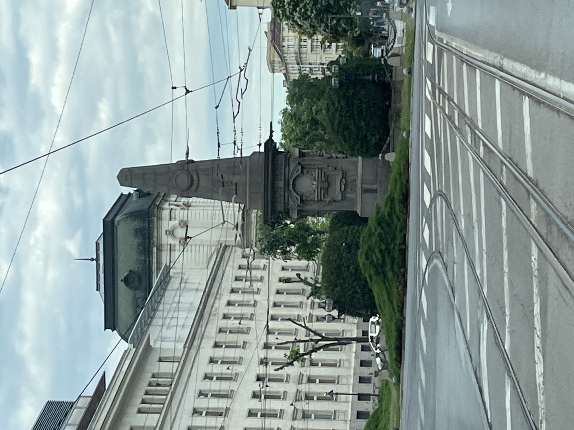In this scene, a monument stands prominently in the foreground, facing a street with trolley tracks and crosswalks. The monument is an obelisk made of dark stone, with intricate carvings on its base. Behind it, a large building with a light-colored facade and many windows is visible, along with lush green trees and bushes. Further in the background, several other buildings and a cloudy sky complete the urban landscape. A white car is parked on the left side of the image. The scene appears to be a city intersection on an overcast day.