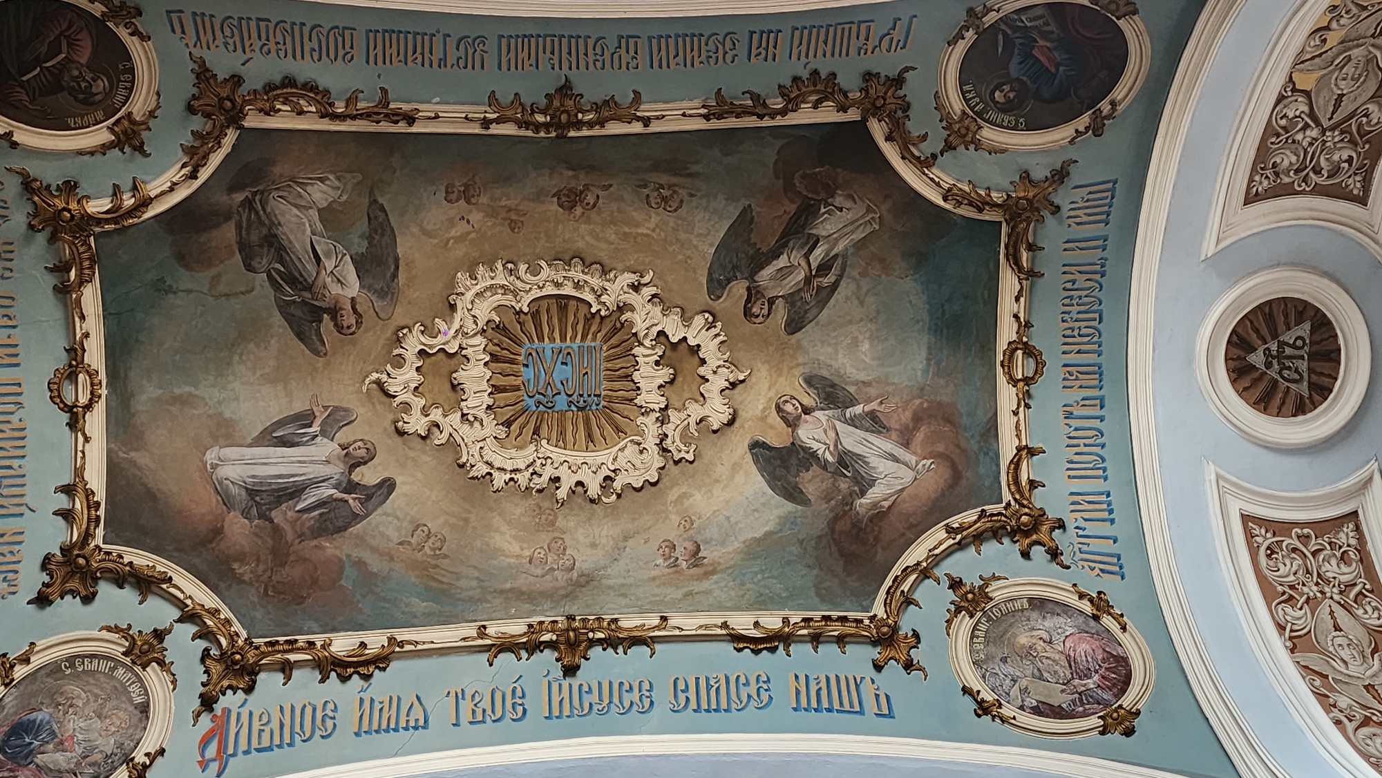 This is a photograph of a painted ceiling, likely within a church. The ceiling features a rectangular central panel with a depiction of angels in a cloudy, ethereal setting. In the center of the panel is a sunburst design with Cyrillic letters inside. The surrounding frame is decorated with ornate, gold-colored scrollwork.
The ceiling outside the central panel is a light blue color. Along the edges of the central panel, there are inscriptions in Cyrillic script in blue lettering. Round medallions containing images of saints and religious symbols are set into the blue background, also framed with gold ornamentation. The scene appears to be located in the city of Unknown, Unknown.